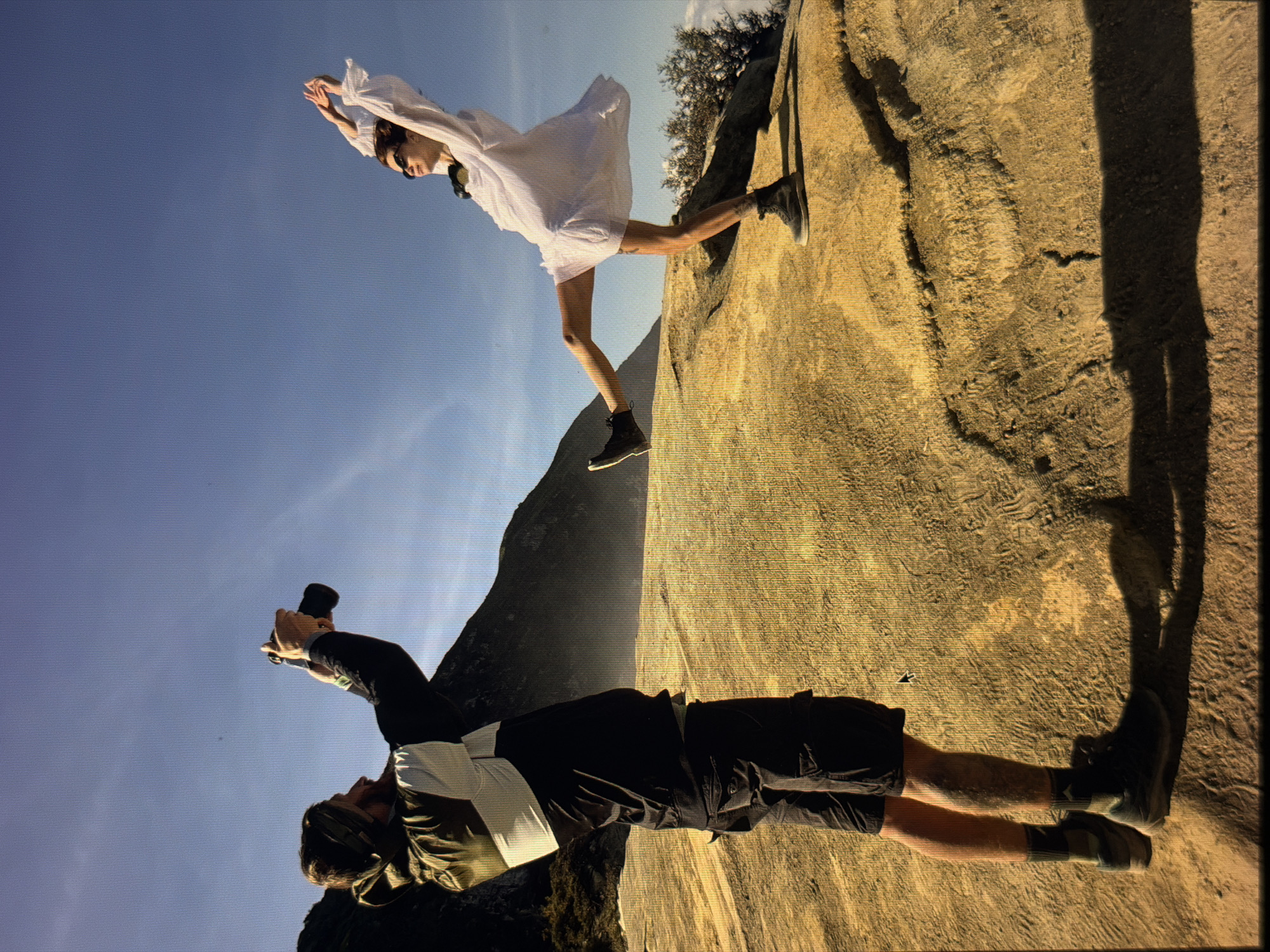 A photograph displays an illusion. A man is lying on a rough, tan-colored rock formation, appearing as if he's scaling a cliff face. He's dressed in dark shorts, a dark shirt with a light-colored vest, and boots. He is holding a camera, pointed upwards.
Above him, a woman in a white dress appears to be floating or jumping against a clear blue sky. She has on sunglasses and dark boots. She's positioned against the sky, but with a part of the rock formation right behind her.
The actual location is likely not in the city of Đà Nẵng, Vietnam, but is an optical illusion created by photographing the scene on its side. The rock formation serves as the "ground" for the woman, making it appear as though she is jumping off a cliff. A darker, distant mountain is also visible in the background, contributing to the illusion of depth and height.