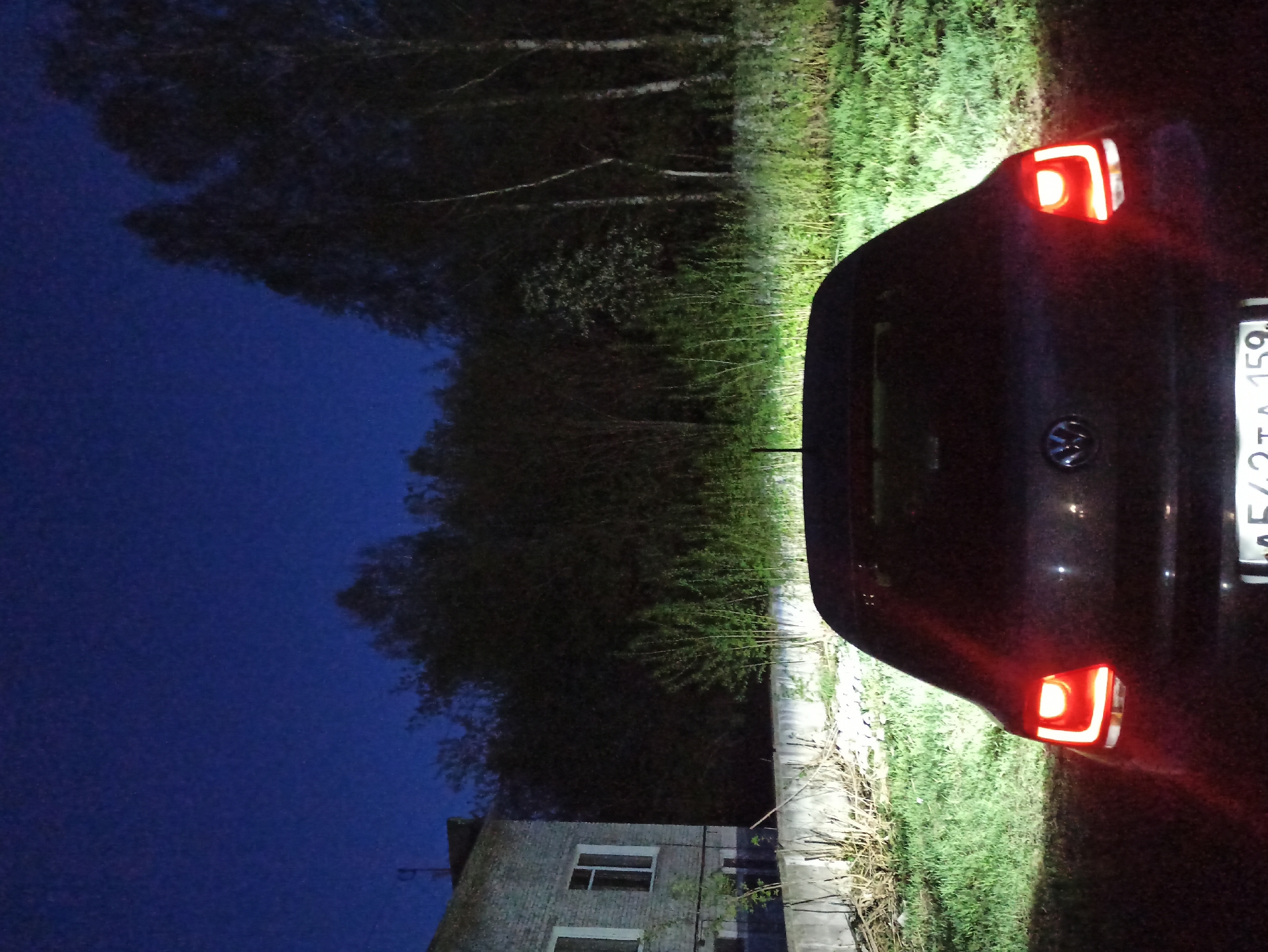 Here's a description of the image:
The image shows the rear of a dark-colored car parked at night. The car's taillights are illuminated. The license plate is partially visible and appears to be a Russian license plate. Behind the car, there's a building with two windows, suggesting it might be a house or a small apartment building. The surrounding environment includes some greenery. The sky is dark, indicating it is either late evening or night.