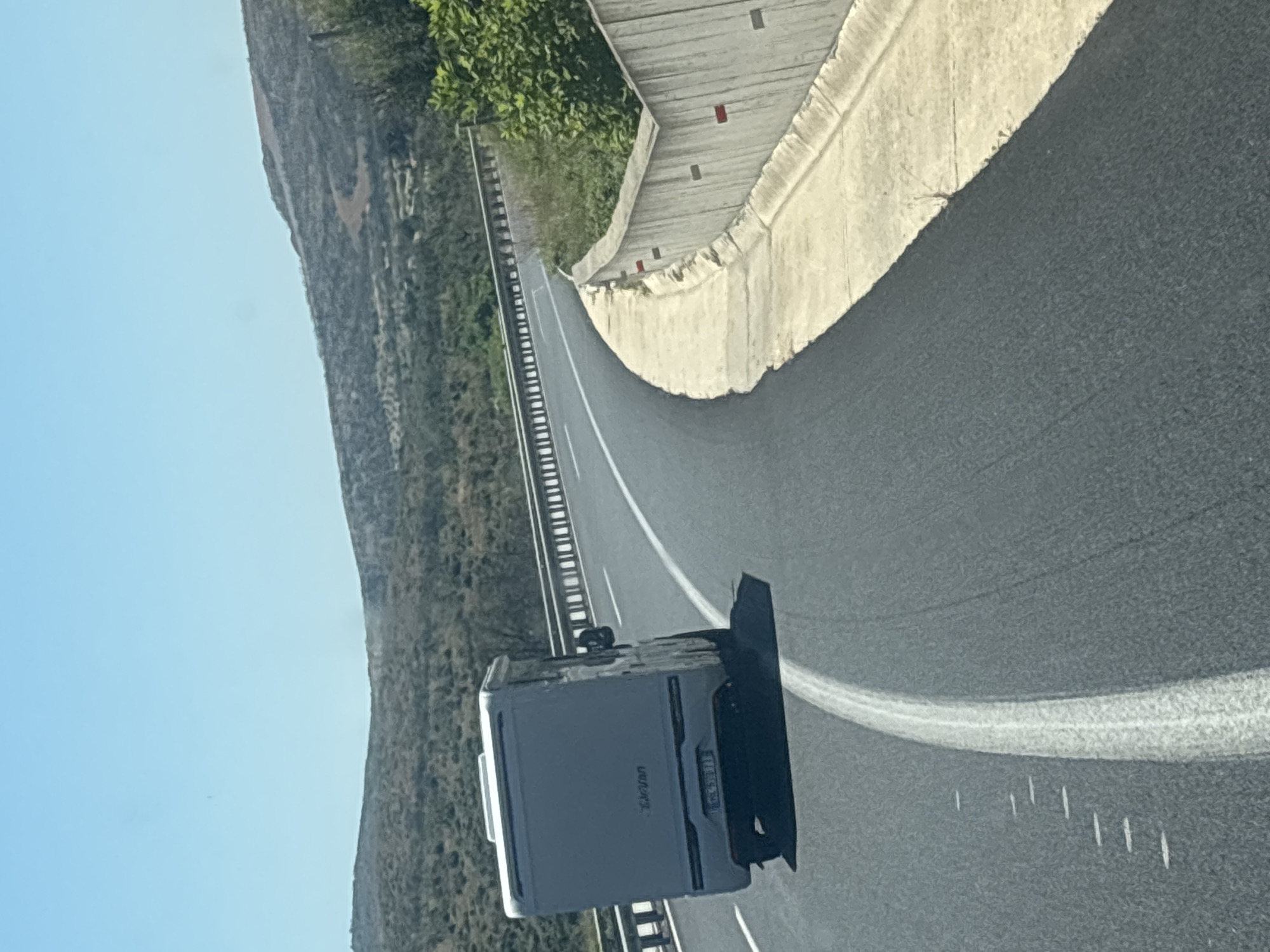 An aerial shot shows a white camper van driving on a highway that curves to the right. The highway is paved with asphalt and has a white lane marker. The camper van is positioned in the left lane, with its rear facing the camera. The background consists of rolling, arid hills covered in sparse vegetation, under a clear blue sky. A concrete barrier with red reflectors lines the right side of the highway. The scene is set in Avra, Greece.