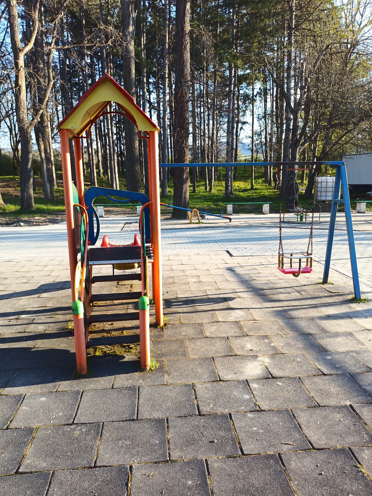 The image captures a playground in Targovishte, Bulgaria, bathed in the soft light of a sunny day. The scene includes a small play structure with orange posts, a yellow roof, and a blue slide. To the right, a blue swing set stands with a single pink and red seat hanging from chains. In the background, tall trees line the perimeter of the playground. The ground is paved with gray square tiles. A seesaw and benches can be observed in the background. A white container is also present on the right. No people are visible.
