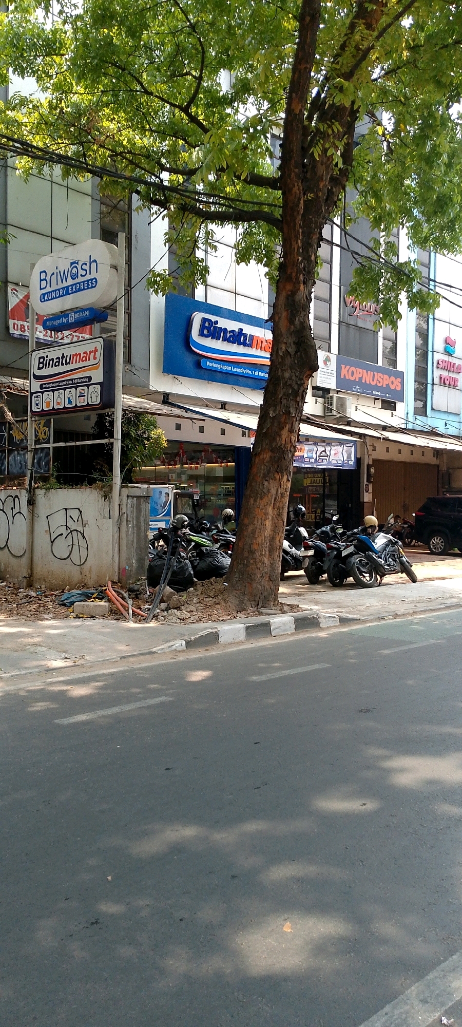 The image shows a street scene in Bandung City, Indonesia, on a bright, sunny day. A large tree with lush green leaves dominates the foreground, its trunk leaning towards the right of the frame. The tree's canopy casts dappled shadows on the asphalt road and the sidewalk.
On the sidewalk, several motorcycles are parked, some with black trash bags piled behind them. There is also debris, including loose soil, leaves, and some orange pipes, near the base of the tree and a concrete wall. The wall has graffiti on it, including large, abstract white shapes.
Behind the sidewalk, there are several businesses with signage. To the left, a sign for "Briwash Laundry Express" is visible, mounted on a pole. Below it is a sign for "Binatumart," featuring icons of laundry services. Further to the right, a prominent blue sign reads "Binatumart" with the tagline "Perlengkapan Laundry No. 1 di Bandung." Adjacent to this is another sign with "KOPNUSPOS" written on it. To the far right, partially obscured by the tree, is a sign for "SHILLA TOUR." The buildings appear to be modern, with glass windows and an awning extending over the storefronts. In the background, a black car is parked on the street. The overall scene suggests a commercial area with a mix of businesses and everyday street life.