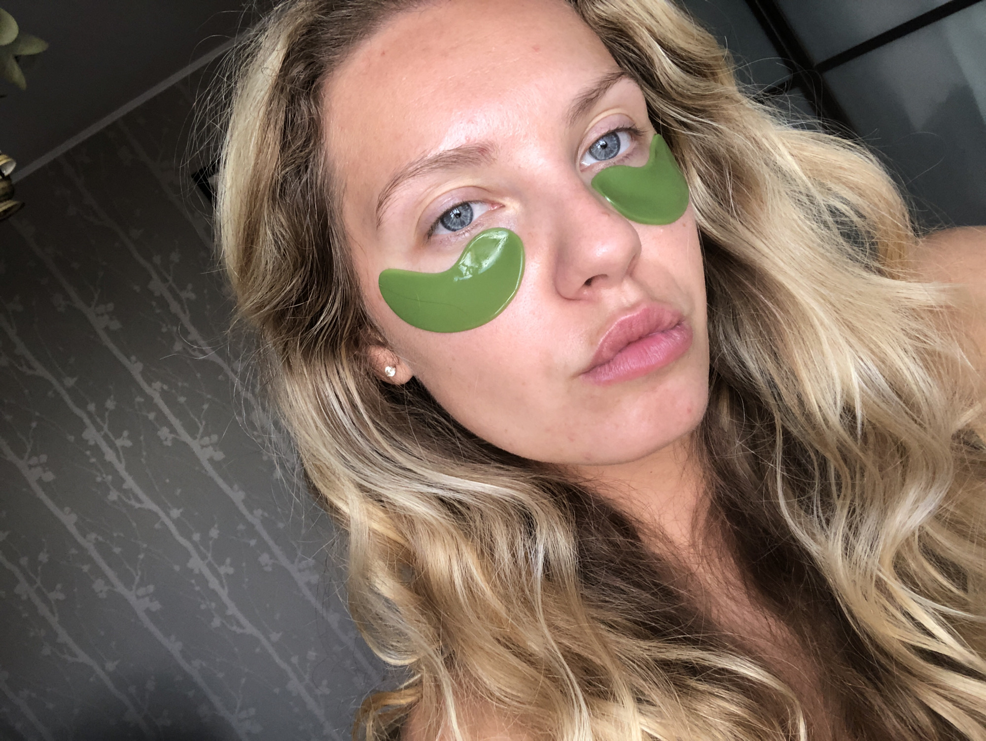 A medium shot, slightly overhead selfie captures a young woman with blonde, wavy hair and blue eyes, wearing green under-eye patches. Her face is turned to the right, with her lips slightly parted and her eyes looking towards the viewer. The background appears to be a dark grey wall with a subtle, repeating pattern of tree branches. The lighting is soft and natural, illuminating her face and highlighting the texture of her hair and the skin. The image seems to be taken indoors, possibly in a bedroom, with no clear indication of the city of Khimki, Russia.