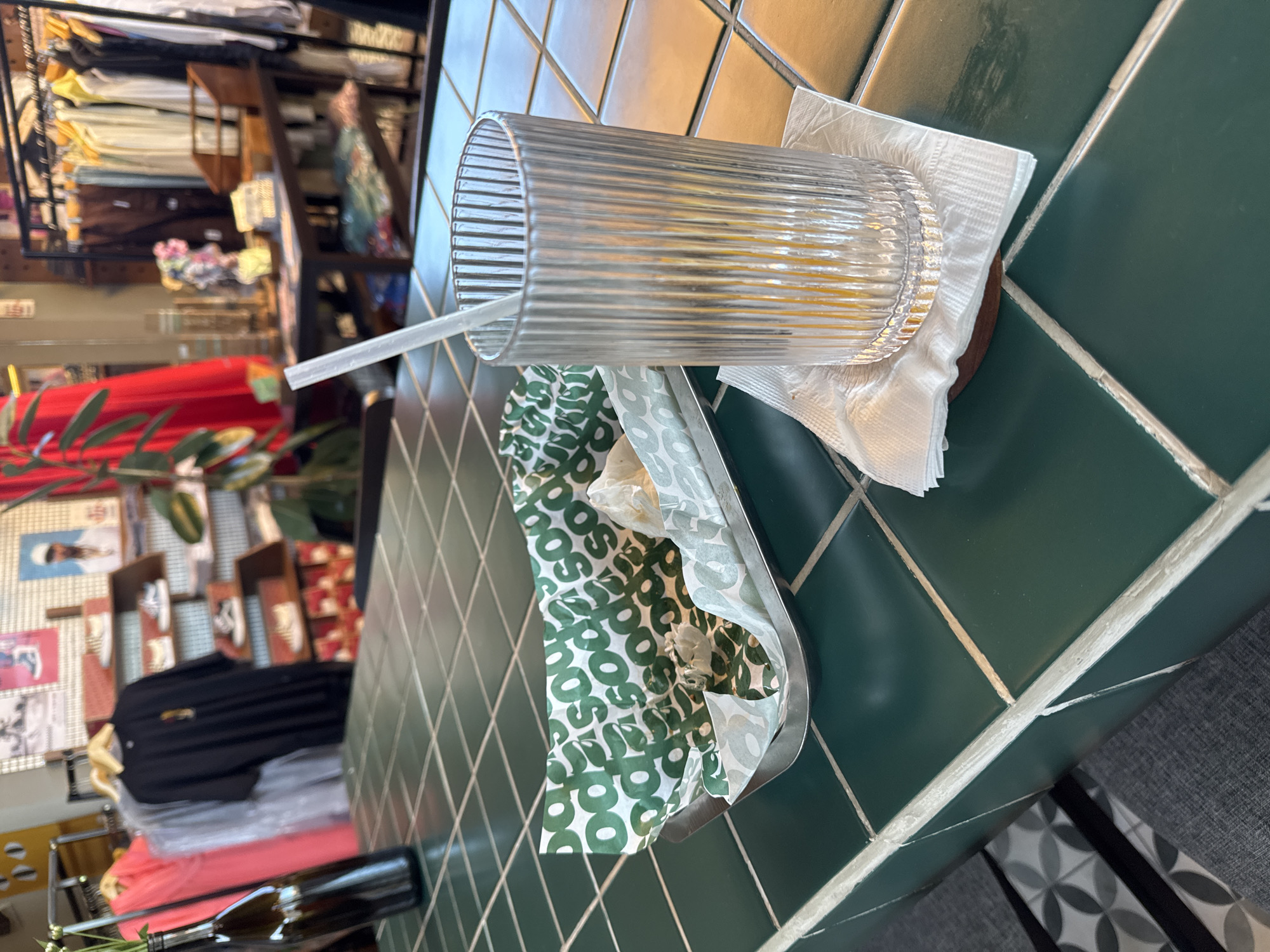 A glass with a straw and some kind of food wrapper sits on a tiled table, with a partial view of clothing racks, shoe displays, and other shop items in the background. The location is likely indoors, possibly a café or restaurant in a retail area. Given the city is Kuta Selatan, Indonesia, the environment would be typically tropical. No people are visible in the immediate frame.