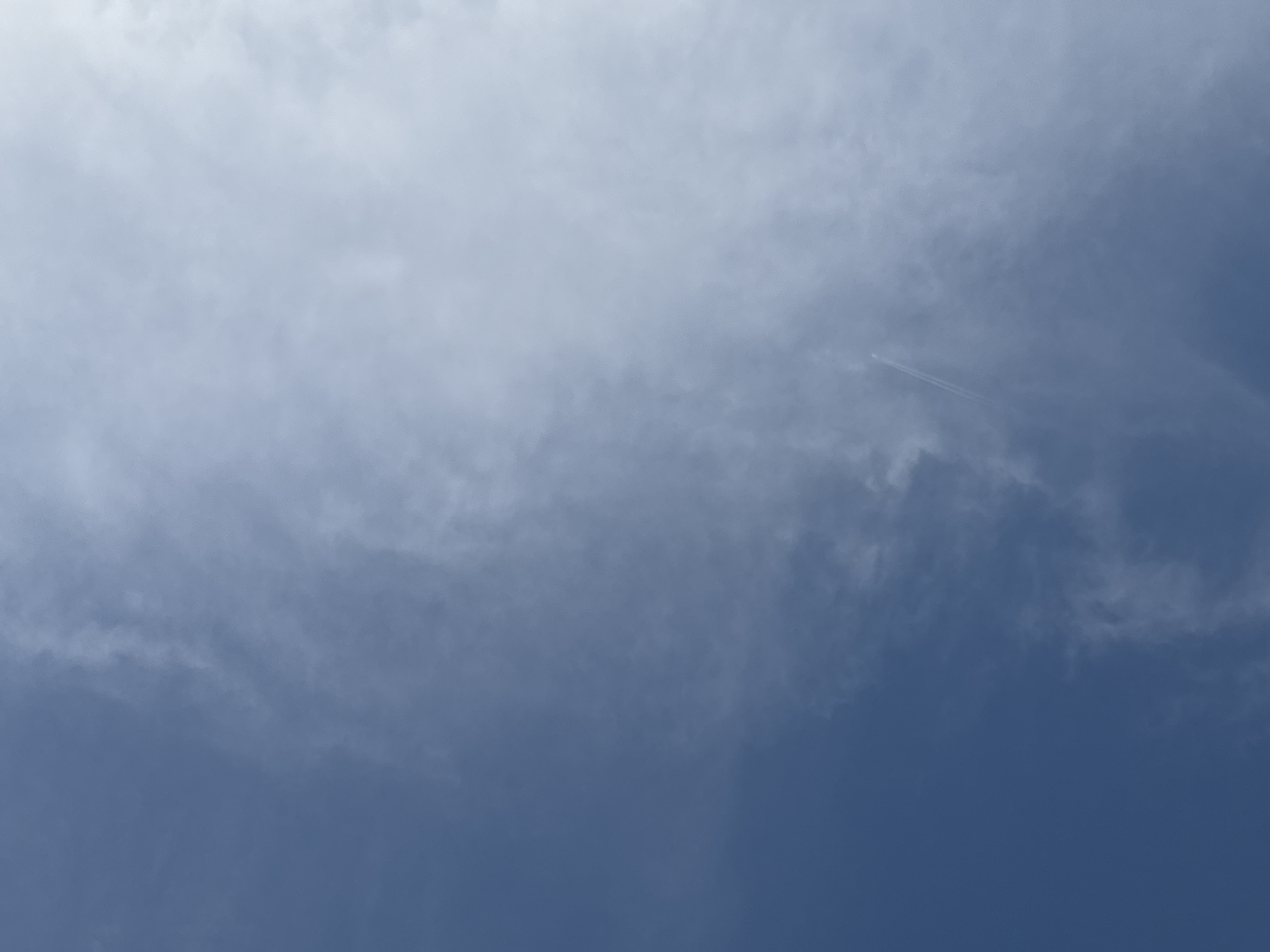 This image shows a bright, partly cloudy blue sky, with the clouds appearing wispy and scattered. A faint contrail from an airplane is visible stretching diagonally across the right side of the frame. The lower left portion of the image is a clear, deep blue sky, while the upper right is dominated by the lighter, diffused light of the clouds. The scene is devoid of any earthly elements, focusing solely on the sky.