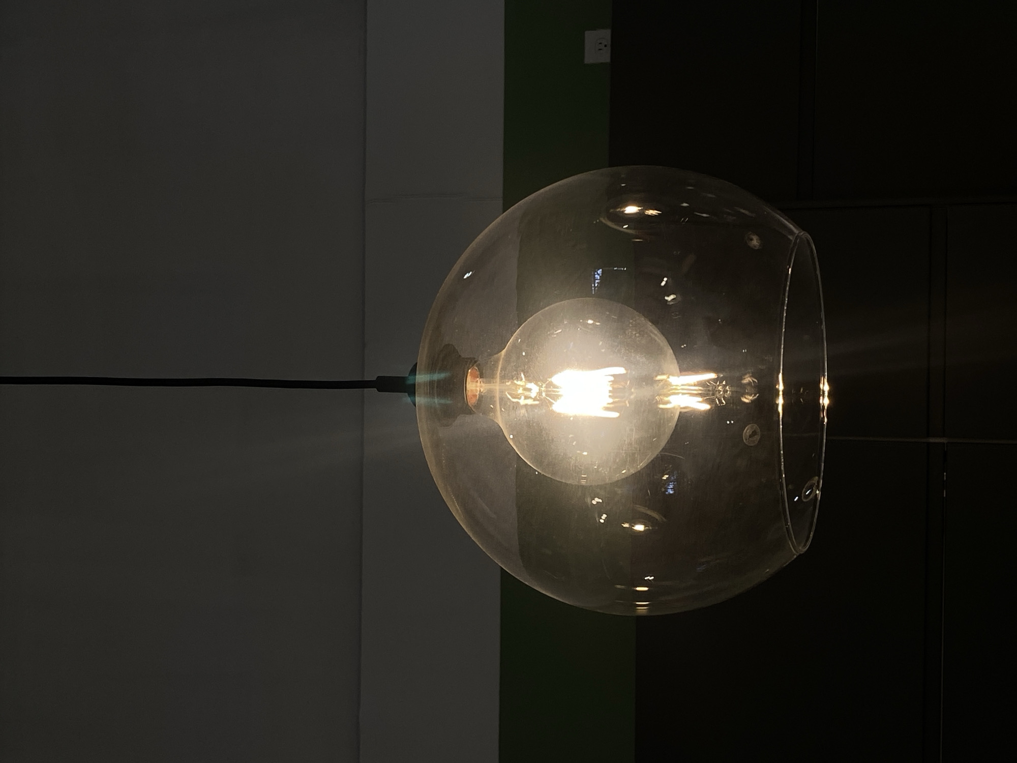 A glass pendant lamp with a visible filament bulb hangs against a backdrop of vertical gray, green, and black panels. The lamp is spherical with an open portion on its right side. A black cord extends horizontally to the left. The scene is dimly lit, emphasizing the bulb's glow. The location is not discernible from the image.