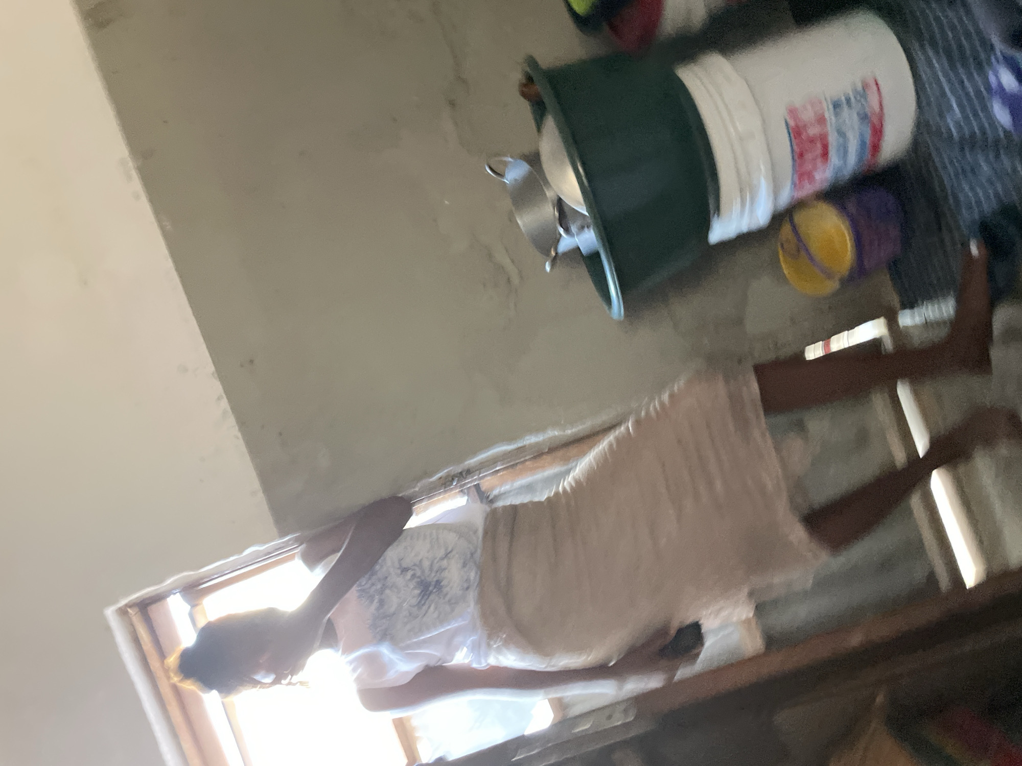 The image depicts an indoor scene, likely a domestic interior, possibly in Jos, Nigeria. A person, appearing to be a young female, stands in the mid-ground, facing towards the left of the frame. She is wearing a white sleeveless top with a blue graphic print and a long, light-colored skirt that reaches her ankles. Her hair is styled up in a bun, and she appears to be holding something to her face with her right hand.
The background features a wall with an off-white, possibly textured or dirty finish. To the left, a window is visible, allowing natural light to enter the room, creating a bright area around the person. There is some furniture or structural element with a dark wooden finish near the window.
In the right mid-ground, several objects are arranged on the floor, including a large green basin filled with metal pots and pans. Beside the basin is a white bucket with some red and blue markings, and to its left, two smaller buckets, one yellow and one purple. The floor appears to be covered with a striped mat or fabric. The overall lighting suggests it is daytime.