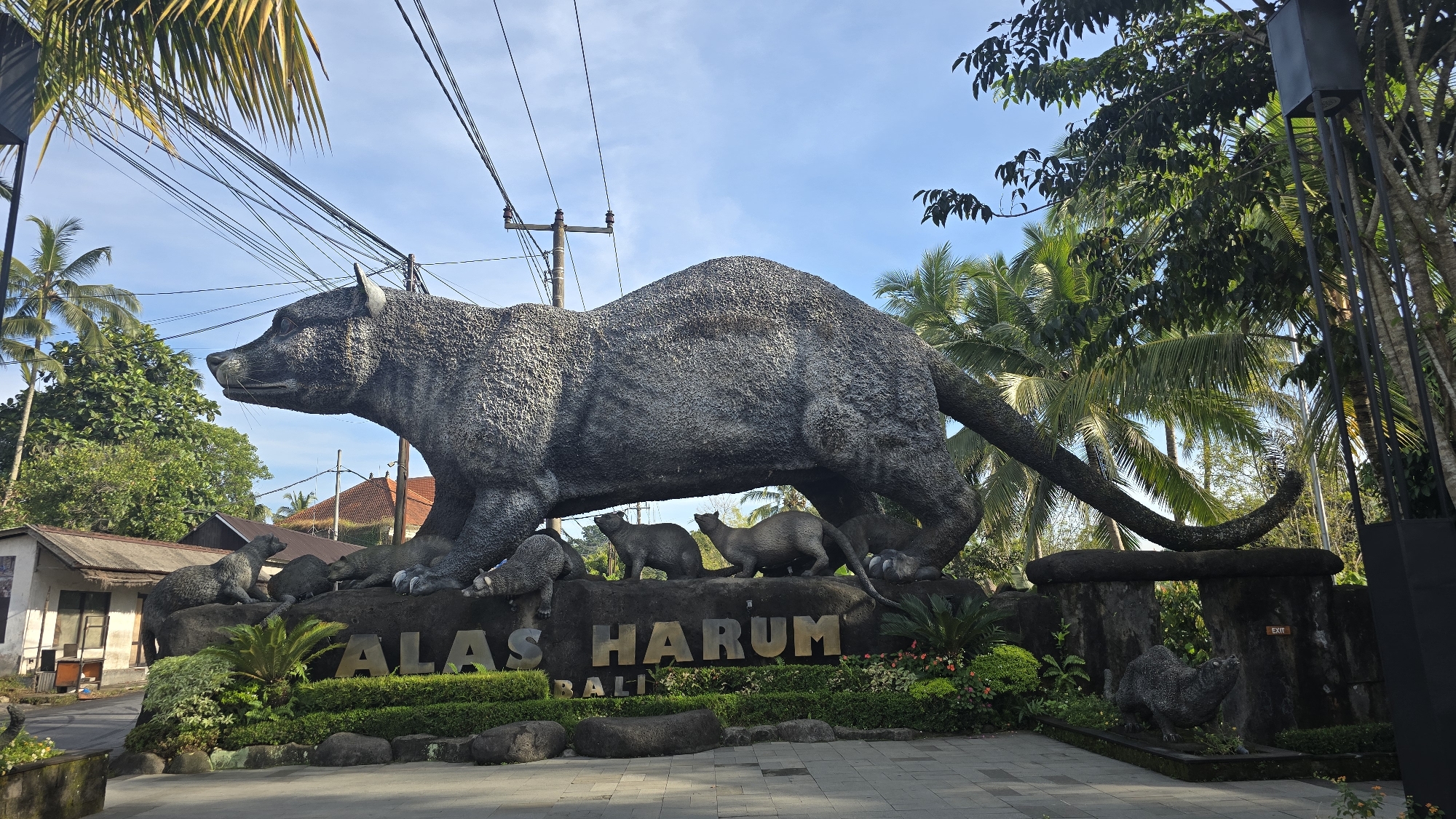 The image shows a large, grey, textured statue of a civet cat on a stone platform, likely located in Tegallalang, Indonesia. Several smaller civet cat statues surround the base of the larger statue, which also features the words "ALAS HARUM BALI" in gold lettering. The statue appears to be an entrance marker or landmark for a location called "Alas Harum Bali." The environment includes tropical trees, foliage, power lines, and a building in the background. The sky is partly cloudy. The statue and surrounding vegetation are well-maintained, suggesting a tourist or public attraction.