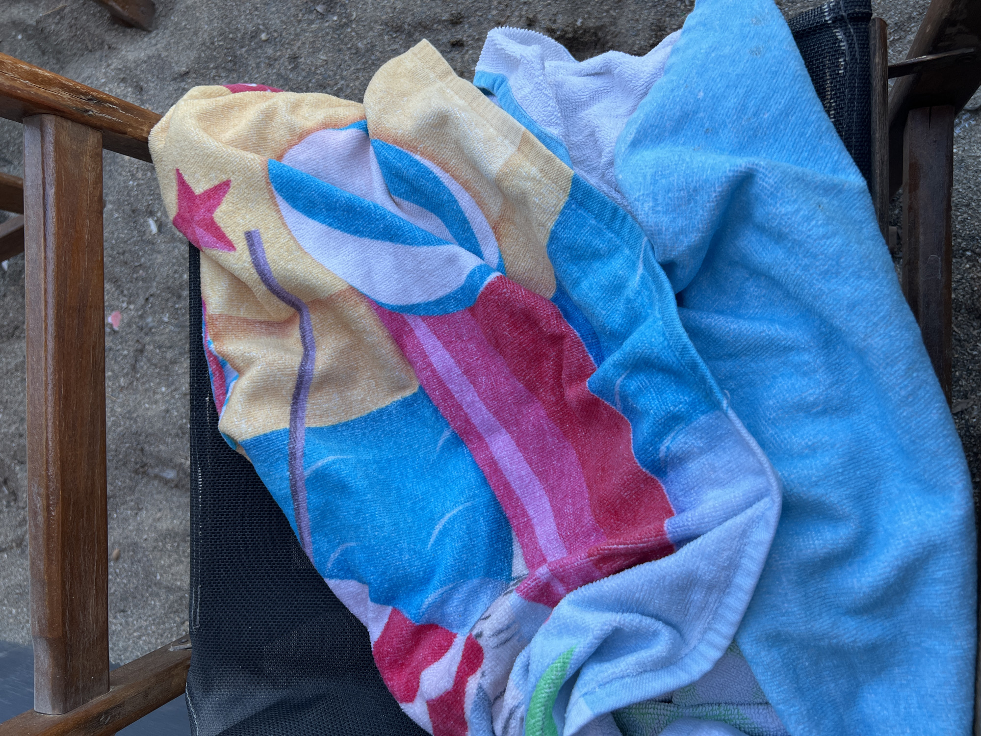 A close-up, overhead shot shows a pile of colorful towels draped over a dark mesh fabric, which is part of a beach chair. The towels are a mix of bright colors and patterns, including pink stars, stripes of red, pink, blue, yellow, and white, and some indistinct circular motifs. The beach chair appears to be made of dark wood and is positioned on a sandy surface. The sandy ground suggests the towels are at a beach or outdoor recreational area. The specific location in Eretria Municipal Unit, Greece cannot be determined from the visual information provided.