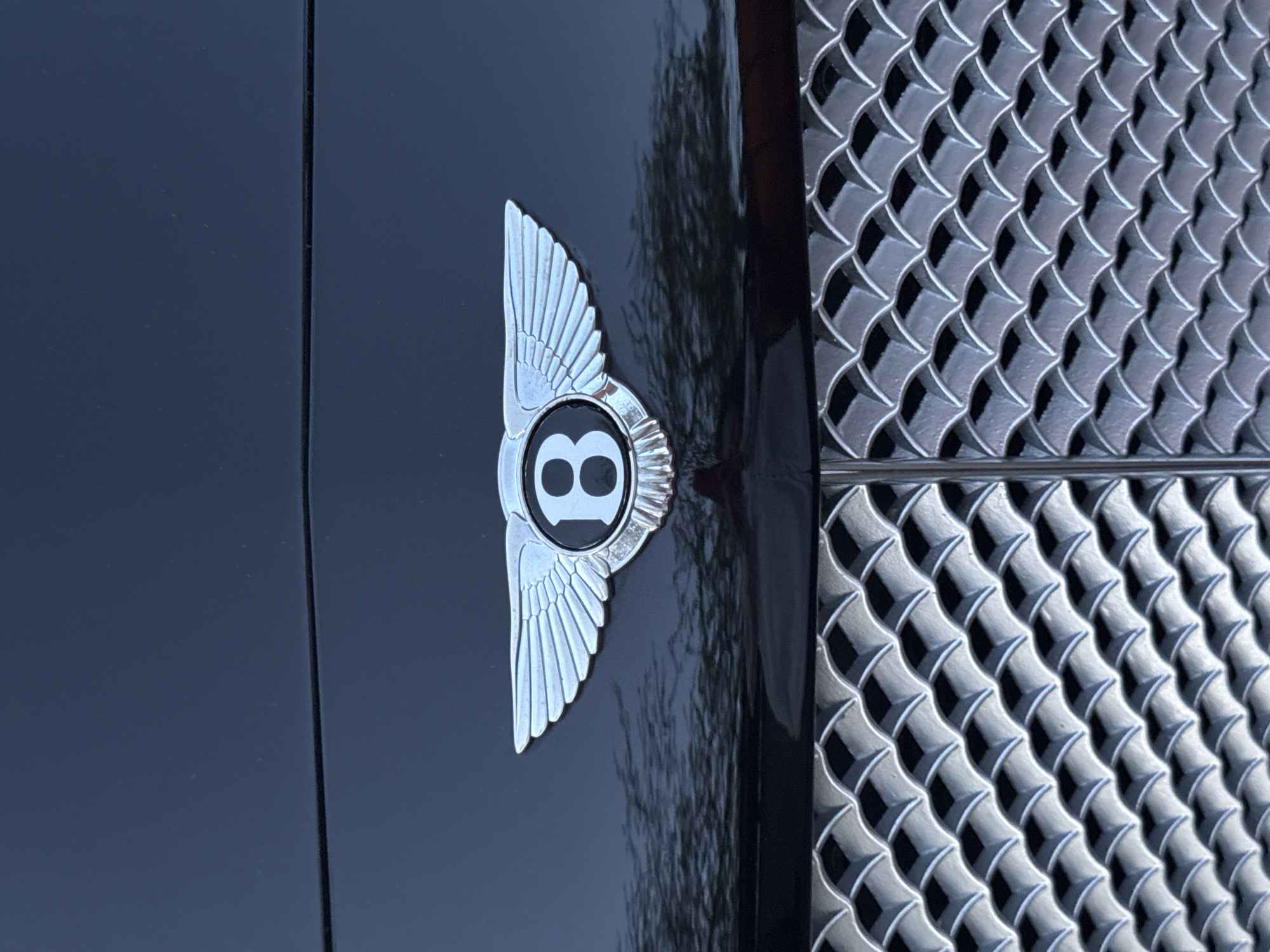 The image shows a close-up of a Bentley car's emblem and grille. The emblem, featuring a winged "B" logo, is attached to a dark-colored, possibly black, panel of the car's body. Adjacent to the emblem is a silver-colored grille with a repeating pattern of diamond or scale-like shapes. The environment is not explicitly visible, so it is not possible to determine the location in Chaikovsky, Russia, or any other specific details about the surroundings.