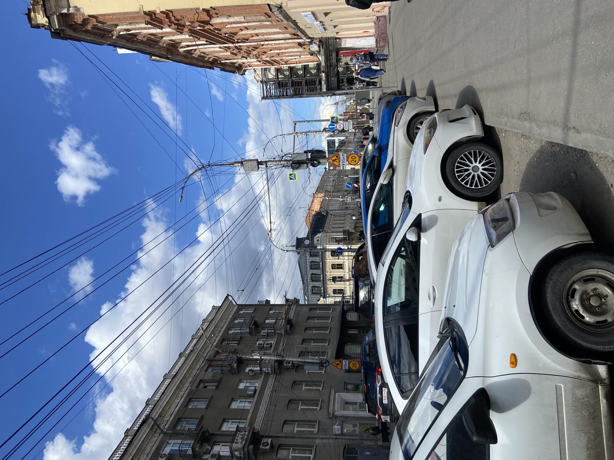 The image captures a vibrant daytime street scene in Saratov, Russia, under a bright blue sky with scattered white clouds. The perspective looks down a city street, flanked by multi-story buildings and a row of parked cars on the right.
On the right side of the street, several cars are parked bumper-to-bumper. The vehicles closest to the viewer are white, while a blue car is visible further down the line. One white car's wheel with a black and silver rim is prominently featured, and another car's simple steel wheel is visible. A license plate "К 848 ТР 64" is visible on one of the cars, with "64" being the region code for Saratov Oblast.
A utility pole dominates the middle ground, laden with a dense network of overhead wires that crisscross the sky. Attached to this pole are various traffic signs: a yellow circular sign indicating a speed limit of "20", several yellow triangular warning signs, and blue circular mandatory instruction signs. One blue sign features "3.5т" (3.5 tons), and another, partially obscured, reads "СТОП" (STOP) in Cyrillic. A rectangular white sign with "495м" (495 meters) in Cyrillic is also present.
On the left side of the street, a facade of a gray multi-story building with numerous windows and exterior air conditioning units is visible. Further down the street, buildings with more ornate, traditional architecture, including brickwork and decorative elements, line the street. In the distance, a building with a clock tower is visible. People can be seen walking on the sidewalks on both sides of the street, adding to the urban activity. The scene is brightly lit by the sun, suggesting clear weather.