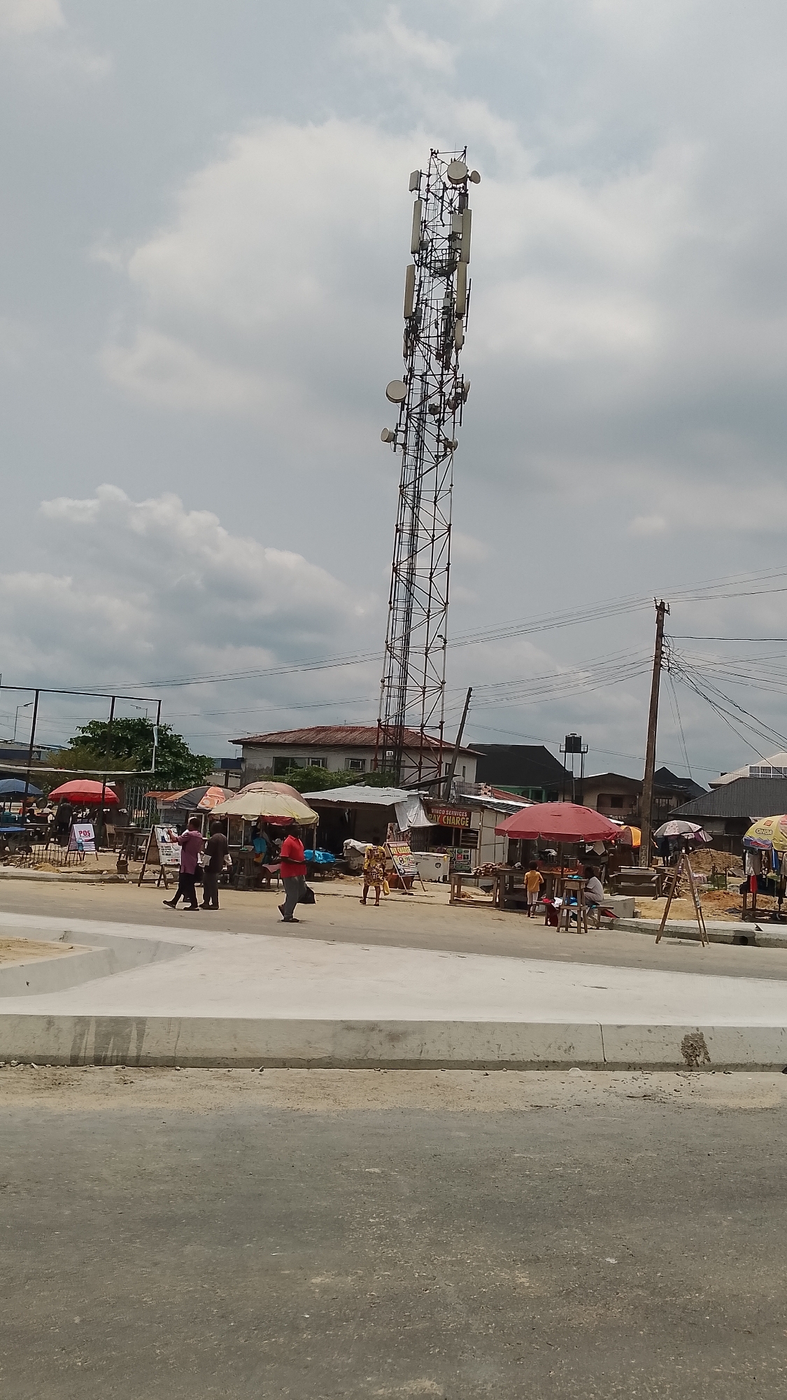 The image depicts a scene in Effurun, Nigeria. A tall, gray telecommunications tower dominates the view, rising high above the low-rise buildings and market stalls that line the street. Several people are walking across what appears to be a newly constructed or renovated section of pavement. Market stalls with colorful umbrellas and goods are visible in the background, suggesting a commercial or trading area. The sky is overcast with gray clouds. Overhead power lines run parallel to the street.