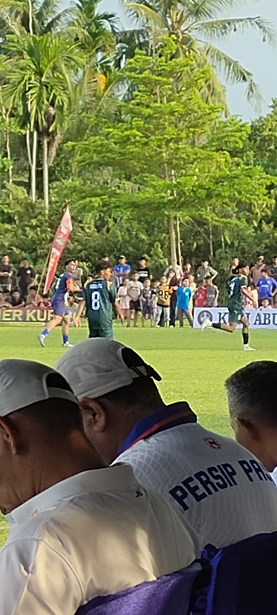 A group of people are gathered in an outdoor setting, likely a sports event, on a grassy field. In the foreground, two individuals wearing white caps are partially visible, facing away from the camera. One of them has on a white shirt with "PERSIP PAR" printed on the back. Behind them, several figures in athletic wear are on the field. One person in a green jersey with the number "8" is prominently featured. A red flag with text is planted on the left side of the field. In the background, a crowd of spectators watches the event, with lush green trees and palm trees forming a natural backdrop. The sky appears to be a light blue, suggesting daylight. The overall scene depicts a lively atmosphere at what appears to be a soccer match, possibly in Keudesimpangempat, Indonesia, given the context provided, as indicated by the text on the jersey possibly referencing a local club.