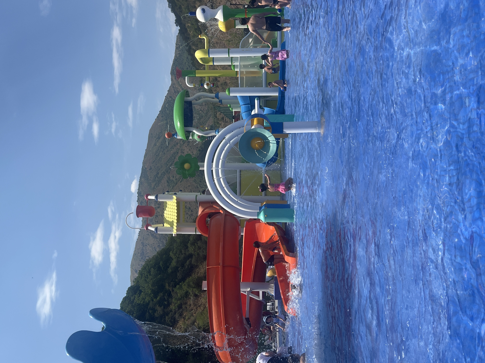 In the city of Khumsan, Uzbekistan, a bright, sunny day is depicted at a vibrant water park. The foreground is dominated by a large, clear blue swimming pool where several children are actively playing and splashing. To the left, a large blue water slide is partially visible, with water cascading down from it. In the center and to the right, a multi-colored water play structure stands tall, featuring various slides, spouts, and interactive elements. Red and orange slides are prominent, with children utilizing them. The background consists of lush green, tree-covered hills and mountains under a clear blue sky with scattered white clouds, suggesting a natural, scenic setting for the amusement park.