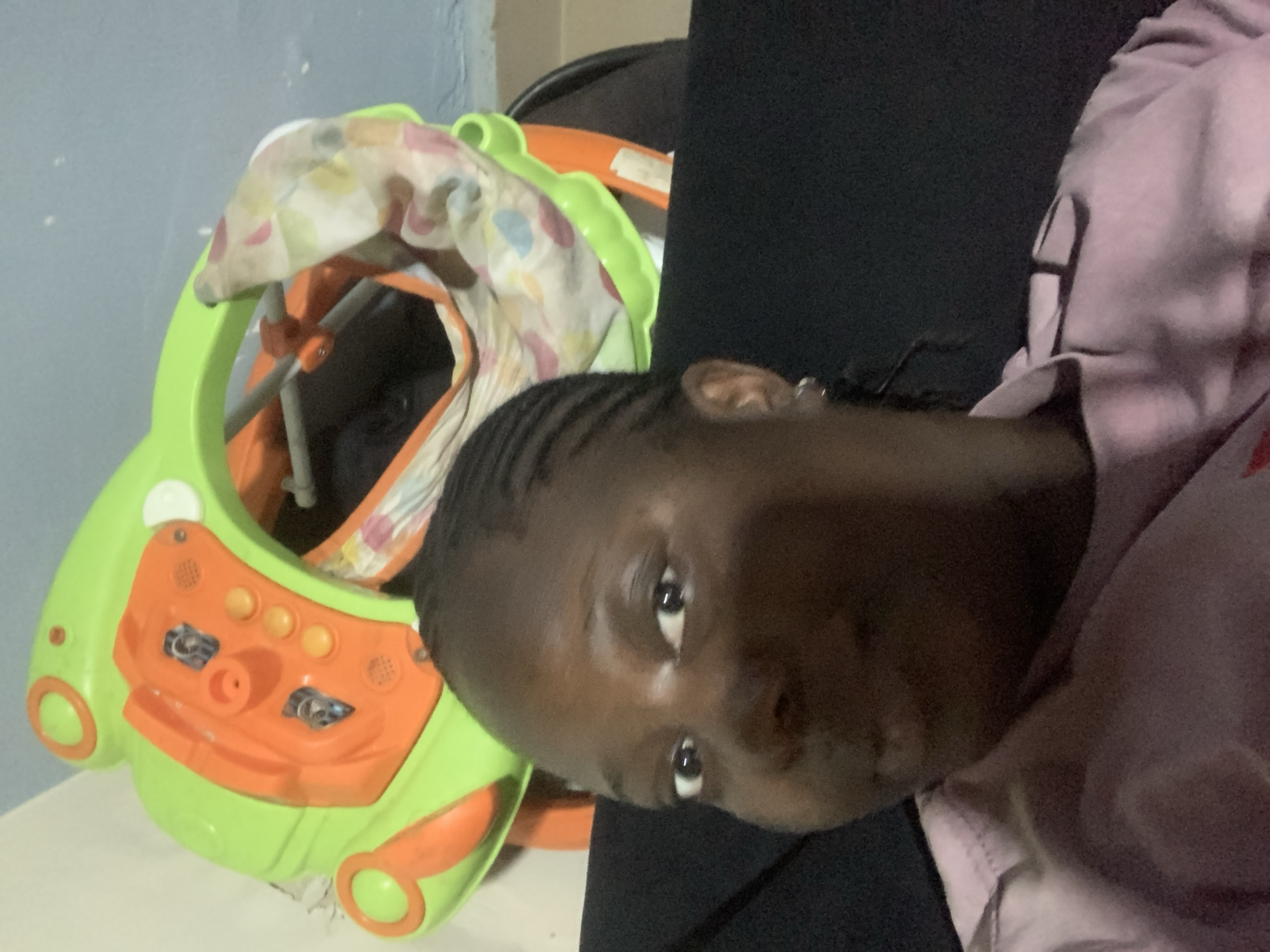 A medium shot captured from a low angle shows a woman with dark skin and braided hair, wearing a pink shirt. She is seated in front of a baby walker. The baby walker is bright green and orange and has a colorful fabric seat with a polka dot pattern. The woman is looking directly at the camera with a neutral expression. The background consists of a light-colored wall and a blue wall, with the baby walker positioned in between.