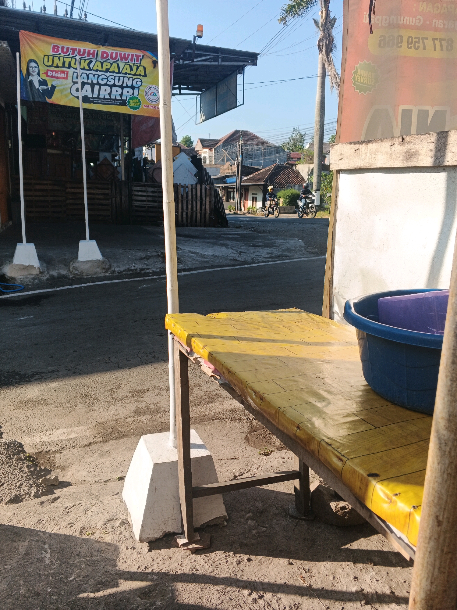 The image depicts a street scene in Semarang, Indonesia. In the foreground, a long, narrow yellow-covered bench with a blue basin containing a purple object rests on a concrete surface. To the left, a banner on a stand reads "BUTUH DUWIT UNTUK APA AJA DISINI LANGSUNG CAIRRR!". Behind this, wooden pallets are stacked. In the background, two people on motorcycles are riding down the street. Further back, houses with tiled roofs and a tall palm tree are visible under a clear blue sky. The overall setting suggests a commercial or roadside area in Semarang.