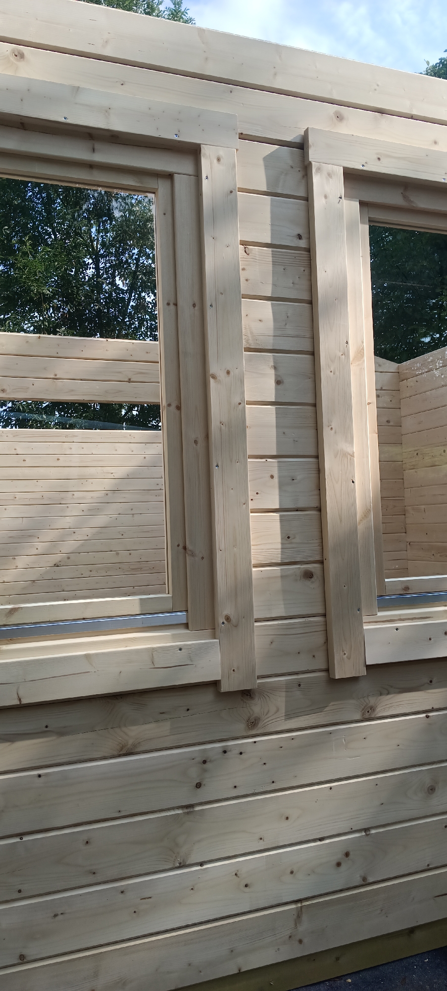 The image shows a partially constructed wooden cabin with several windows. The cabin is made of horizontal wooden planks. There are two visible windows, one on the left and one on the right side of the frame. The windows have wooden frames and appear to be made of glass, reflecting trees and the sky. Vertical wooden beams are attached to the exterior of the cabin, framing the windows and providing structural support. The ground beneath the cabin is not clearly visible, but a dark surface can be seen at the bottom of the frame. The overall impression is of a construction site for a wooden building. The image does not contain any people. The location is specified as Lejšovka, Czechia.