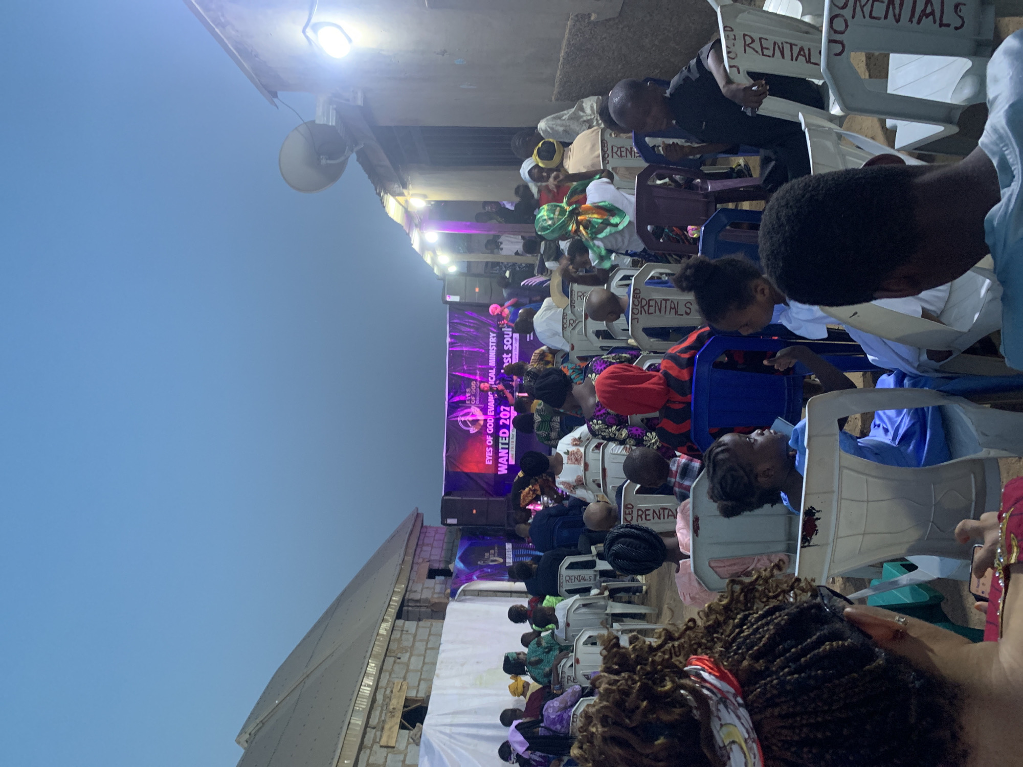 The image shows an outdoor event in Sabojibi, Nigeria. A crowd of people sit in plastic chairs, many marked with "RENTALS," facing a stage. The stage has a banner that reads "EYES OF GOD EVANGICAL MINISTRY" and "WANTED 207." There is a loudspeaker and a street light visible above the crowd. The sky is clear and blue in the background.