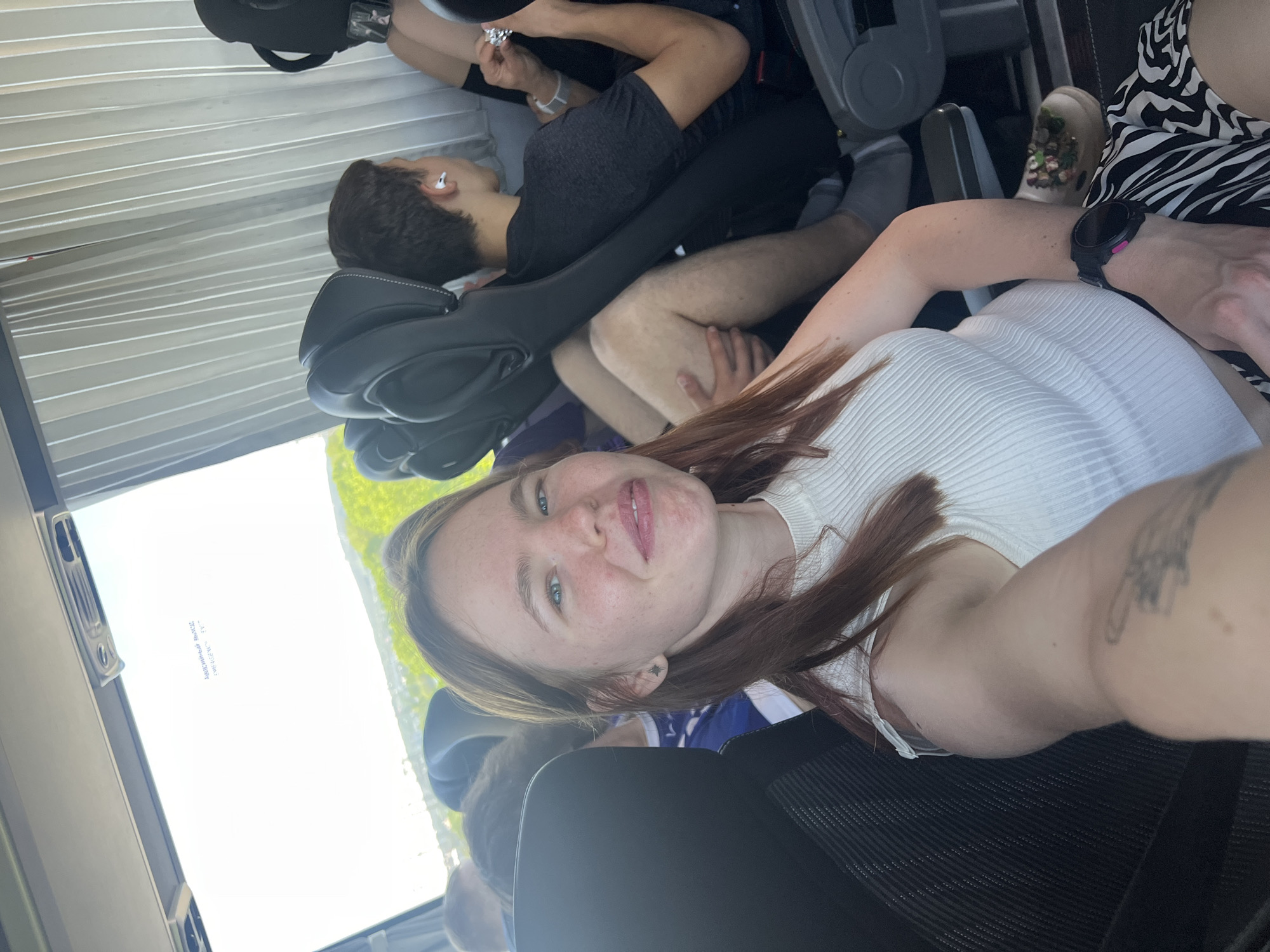 The image shows a woman with red hair taking a selfie indoors. She is wearing a white shirt and a black watch on her wrist. She has a tattoo on her arm. Behind her, there is a man lying down with headphones in his ears. Another person is sitting behind the man. The interior appears to be a bus or a similar vehicle. The presence of a window suggests the location is external. While there is no direct visual evidence to confirm the location as Chaikovsky, Russia, the scene is consistent with being inside a vehicle with passengers.