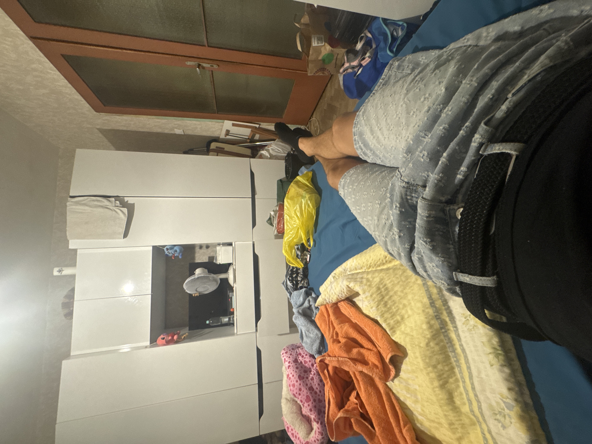 The image shows a bedroom in Sofia, Bulgaria. The room appears somewhat cluttered. On the left side of the image are large white wardrobes. In the center, on a blue bed, are several folded towels, one orange and one yellow, and a pink and white patterned dog bed. A person, whose legs and torso are visible, is lying on the bed. They are wearing light blue denim shorts with a white pattern and black socks. A black belt is visible around their waist. To the right of the bed is a wooden door with frosted glass panels, and beyond that, a glimpse of another room. In the background, near the wardrobes, a small white fan is visible on a shelf. There are also various bags and objects scattered around the room, including a bright yellow plastic bag. The lighting suggests it is either evening or a dimly lit room.