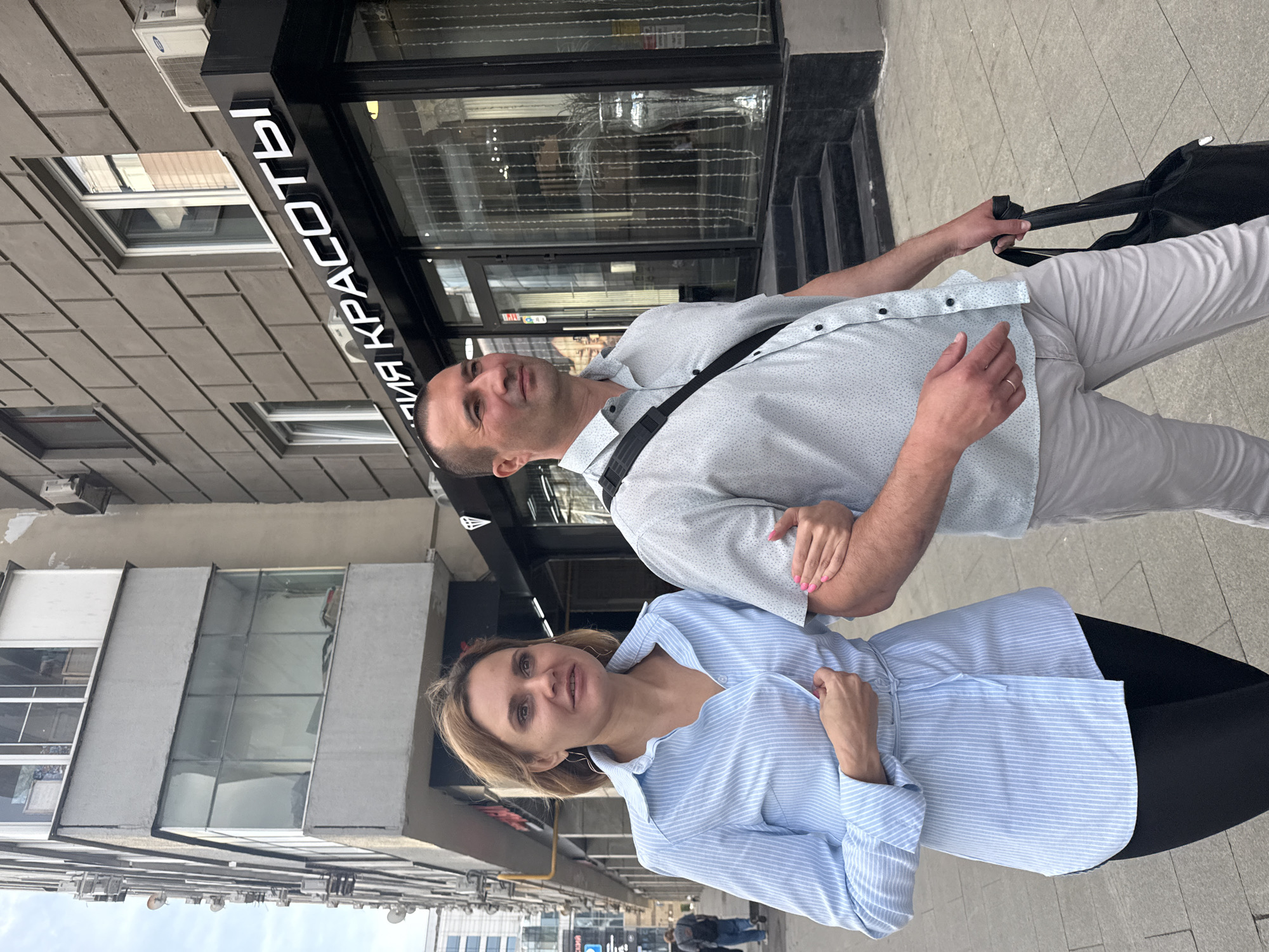 In this vertical, eye-level shot, a couple stands on a sidewalk in front of a building with a black facade and large windows. The building's signage reads "ЛИЯ КРАСОТЫ" in white, raised letters. The man on the right is wearing a light blue, short-sleeved button-down shirt with a subtle dot pattern and light grey pants. He has a black bag slung across his chest and is holding the strap with his right hand. A silver ring is visible on his left ring finger. The woman on the left is wearing a light blue and white striped, long-sleeved button-down shirt. Her hair is blonde and shoulder-length, and she is wearing large hoop earrings. Both individuals appear to be looking at the camera. Their expressions are neutral to slightly smiling. The scene suggests daytime, with overcast or diffused lighting. In the background, a street with parked cars and pedestrians can be seen. The architectural style of the building and the visible text suggest an urban environment in Moscow, Russia.