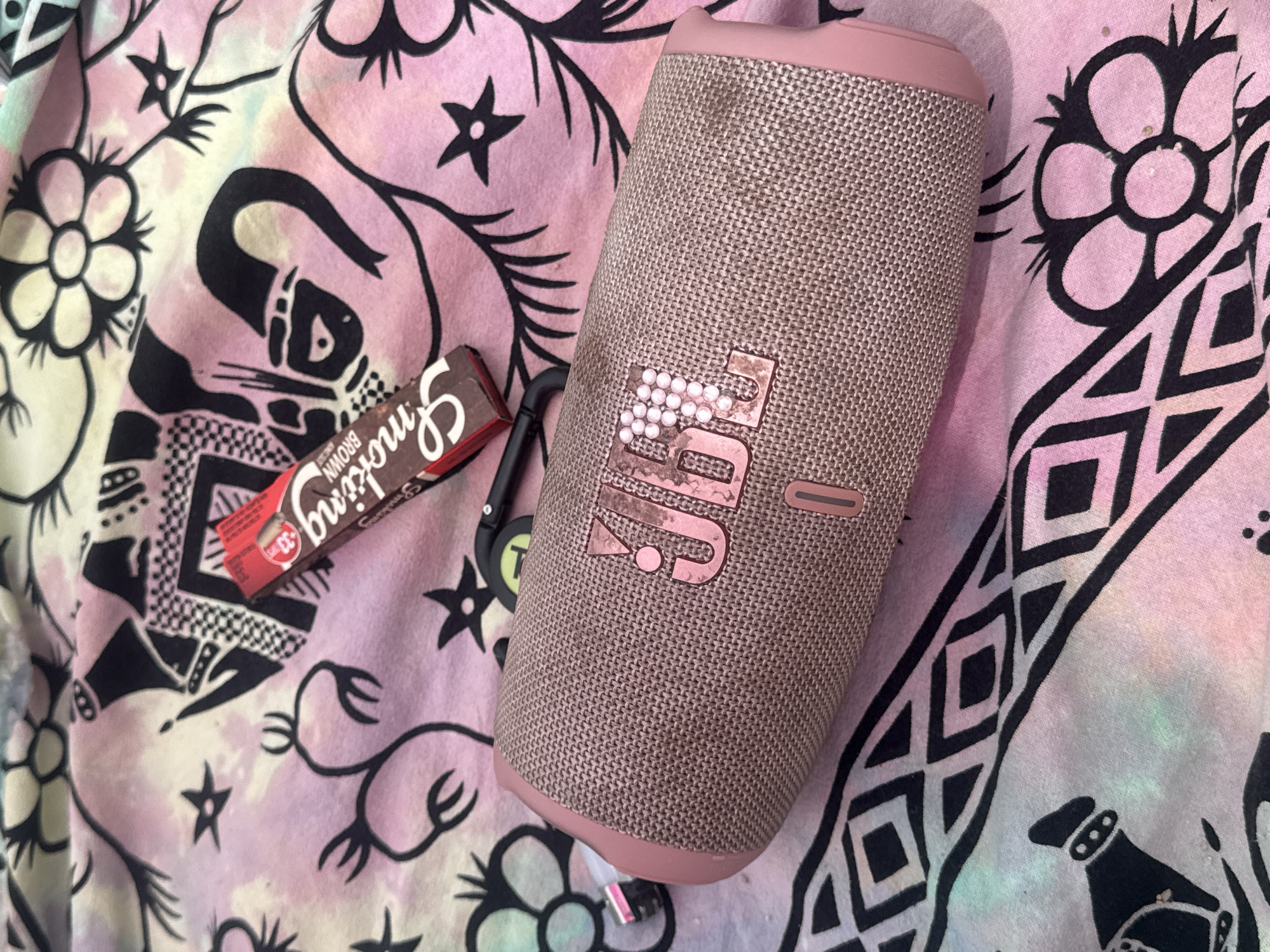 A close-up shot shows a pink JBL portable speaker on a colorful, tie-dyed fabric. The fabric has a black graphic print of stylized flowers, stars, and abstract shapes. A small, red chocolate bar wrapper is positioned above and slightly to the left of the speaker. The speaker is textured and has the JBL logo emblazoned on it with small white beads. A black carabiner clip with a green tag is attached to the side of the speaker. A pink charging port cover is visible on the left end of the speaker. The scene is dimly lit, with a soft focus. The location is not specified in the image.