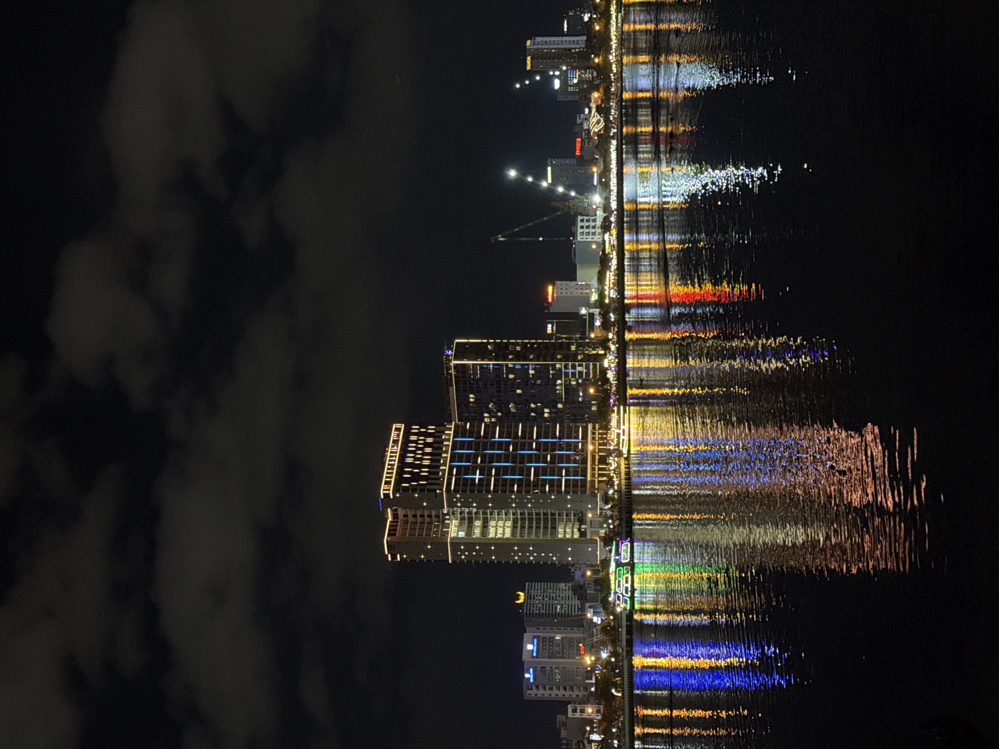 The image depicts a city skyline at night, reflected in a body of water. The perspective is rotated, so the buildings appear to be on the left and the water on the right. The buildings are illuminated with various lights, creating a vibrant display against the dark sky. Some buildings are tall and modern, featuring grid-like patterns of lights, while others are shorter. A prominent crane is visible among the structures. The water's surface is disturbed, causing the lights from the buildings to form elongated, shimmering reflections that stretch across the frame. The reflections showcase a spectrum of colors, including white, yellow, orange, red, green, and blue. The sky above is dark, with some visible clouds, suggesting it's nighttime, and potentially a bit overcast. The scene conveys a sense of urban energy and the beauty of city lights at night, amplified by the mesmerizing reflections. There is no visible text in the image.