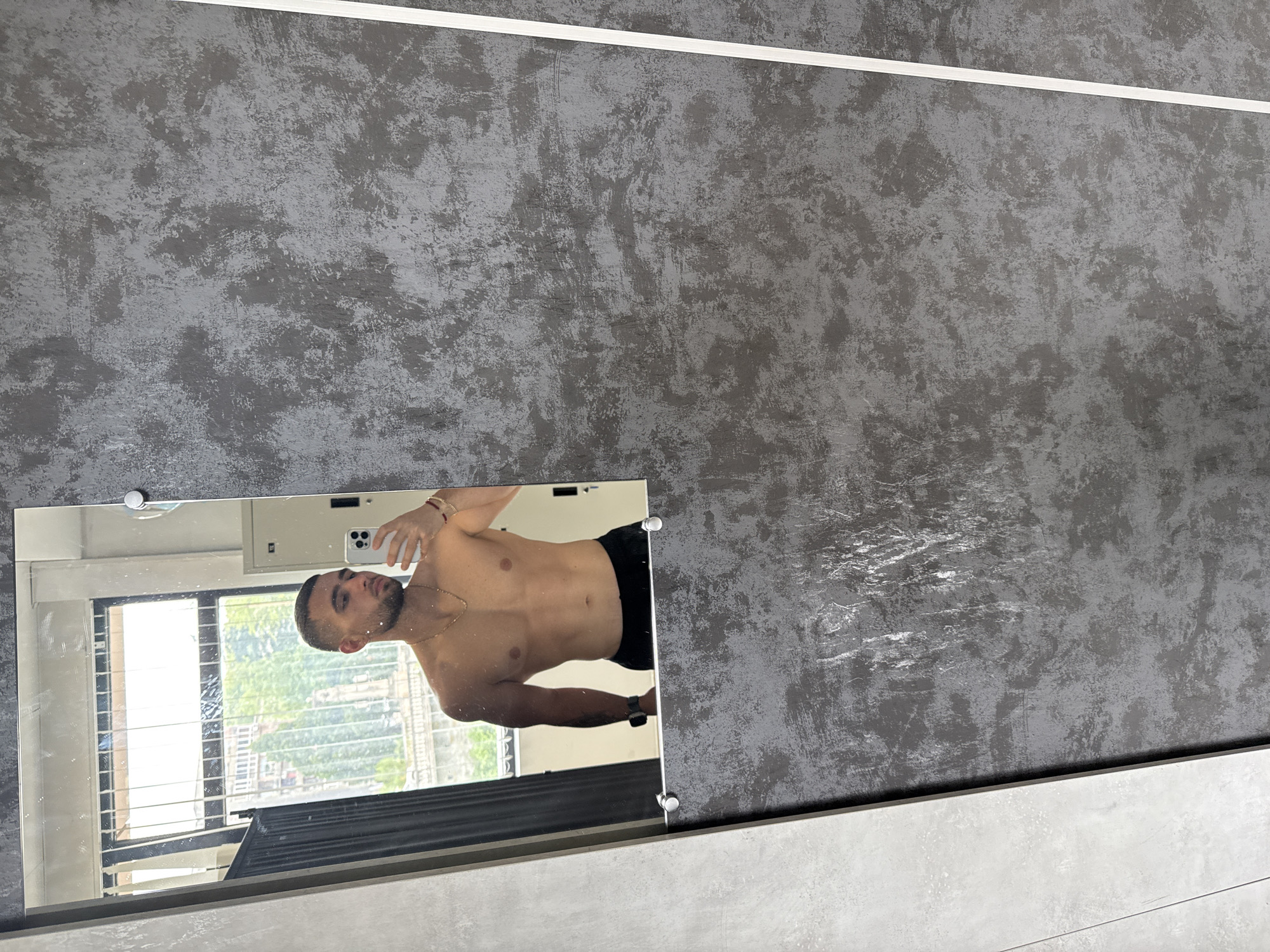 The image shows a man reflected in a mirror, which is mounted on a grey textured wall with a white trim. The reflection reveals a partial view of a window with vertical blinds and a city landscape, possibly Gabrovo, Bulgaria. The man is shirtless and appears to be taking a photo of himself in the mirror with his mobile phone. He is wearing black shorts. The mirror is rectangular with visible mounting hardware.