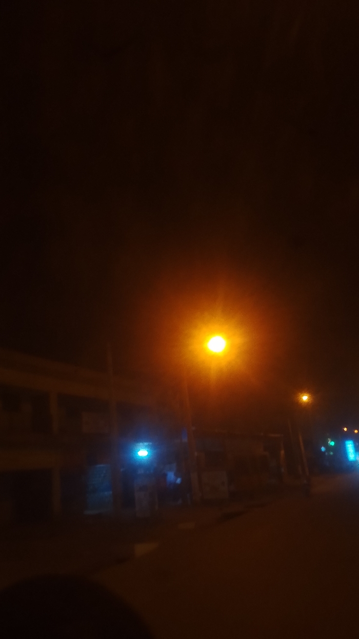 A dark street scene at night features a bright orange streetlight dominating the view. The illumination reveals a low building on the left and several businesses storefronts behind the lamppost. A blue light emanates from one of the storefronts. Further down the street, smaller lights dot the distance. The ground is dark, reflecting some of the orange light. A blurry dark object appears in the bottom left corner, possibly indicating the camera’s lens or the photographer's hand.
