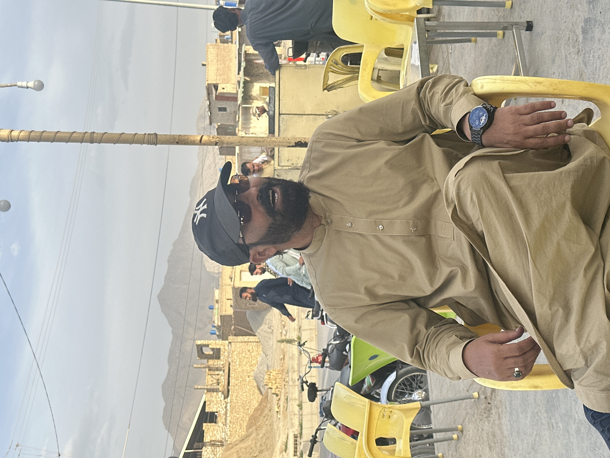 A man wearing a black cap, sunglasses, and a tan shirt is sitting in a yellow chair outdoors. He is wearing a watch on his left wrist and a ring on his right hand. Behind him is a street scene with buildings, motorcycles, and other people. In the background, there are mountains under a cloudy sky. The location appears to be in a built-up area of Quetta City Tehsil, Pakistan.