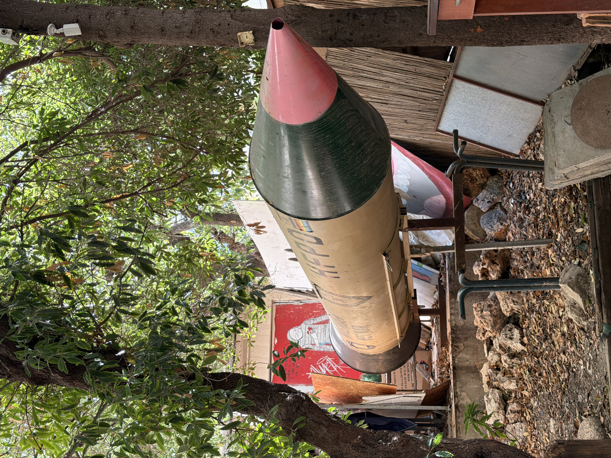 The image depicts an outdoor scene, possibly in Haifa, Israel, featuring a large, cylindrical object resembling a missile or rocket mounted on a stand. The object is painted in a tan color with a dark green section and a pink tip. Text is visible on the side of the object, with the letters "AFPD" clearly legible, and other text is partially obscured.
The missile-like object is positioned in a cluttered area with trees providing overhead canopy and dappled sunlight filtering through the leaves. In the background, there are various objects, including what appears to be a mural or artwork on a wall, depicting a figure with a heart symbol. There are also stacks of wood, scrap metal, rocks, and debris scattered around the scene. A tree trunk is prominent on the right side of the image, with a small white object mounted on it, possibly a sensor or camera.
The ground is covered with fallen leaves and dry foliage, indicating an outdoor environment. The overall impression is one of an artistic installation or a storage area filled with disparate items, set within a natural, albeit somewhat unkempt, environment. There are no visible people in the image.