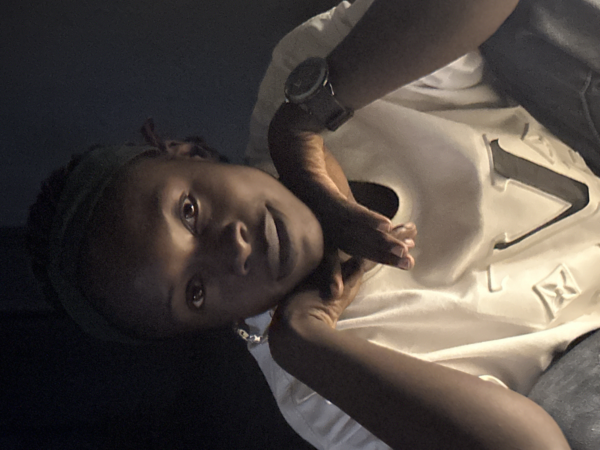 A young Black woman with dark skin and dark hair, wearing a green headscarf and a white t-shirt with a grey design and a dark smartwatch, is pictured in a dimly lit indoor setting, likely a residence in Jos, Nigeria. She is looking directly at the camera with her arms crossed, resting her chin on her hands. The lighting is dramatic, with the subject's face illuminated from the front, creating strong shadows on the left side of her face and on the background, which appears to be a dark wall or curtain. The overall atmosphere is intimate and possibly candid.