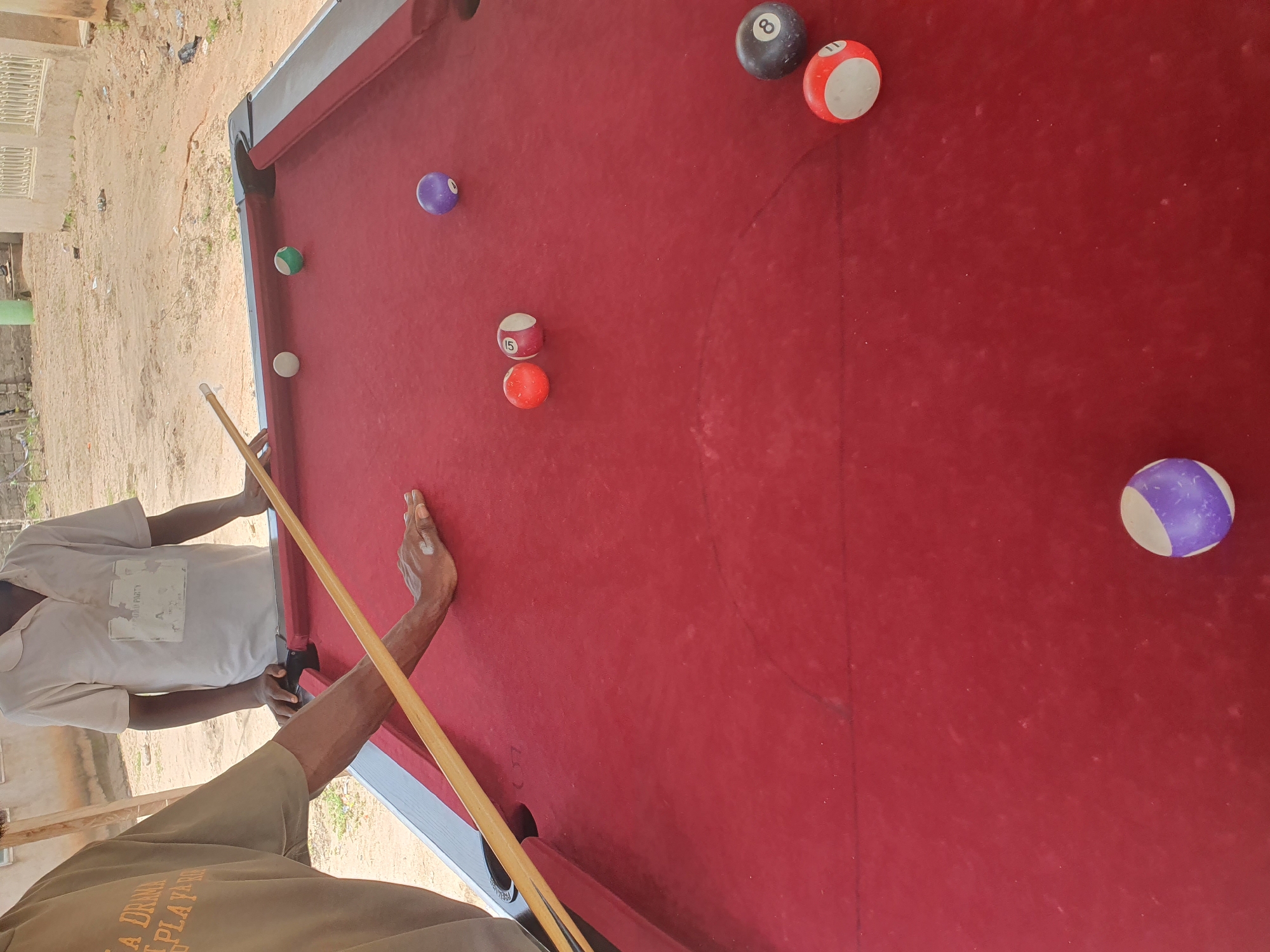 The image depicts an outdoor scene in Lokoja, Nigeria, focusing on a red billiard table with several pool balls. Two individuals are partially visible. One person, wearing a tan t-shirt with text that reads "A DRAMA PLAYARD", is positioned on the left side of the frame, with their right arm resting on the billiard table and holding a pool cue. The other person, standing behind the table to the right, is wearing a white polo shirt with a label that says "TEAM PARTY". The table's surface is red, and the balls present include a purple striped ball, a solid green ball, a white ball, two solid red balls, one with the number 15, another solid red ball with white stripes and the number 11, a solid purple ball, and a black ball with the number 8. The background shows a sandy, unpaved area with buildings and sparse vegetation, indicative of an outdoor setting in Nigeria. The lighting suggests it is daytime.