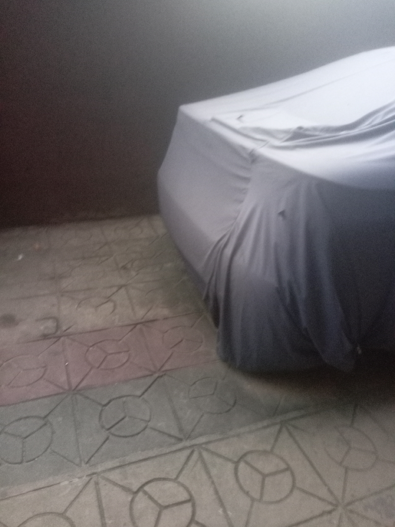 A still, slightly grainy photograph captures a car covered with a light grey protective cover, parked next to a dark, textured wall. The covered vehicle dominates the right side of the frame, its form gently suggested by the fabric's drapes and creases. The wall, appearing dark brown or grey, recedes into the background on the left. The ground in the foreground and beneath the car is paved with distinctive, multi-colored concrete tiles, featuring an intricate geometric pattern of interlocking circles divided into three segments, interspersed with hexagonal and triangular motifs. The tiles exhibit hues of grey, brown, and hints of muted red. The lighting is dim and diffuse, casting no sharp shadows, which could indicate an indoor space like a garage or carport, or an outdoor area under overcast skies. No individuals are present, and no specific activity is taking place, other than the car being stored or parked. The image provides a quiet scene of a vehicle in storage within what appears to be a residential or commercial setting in Iyiowa Odekpa, Nigeria. No text is visible.