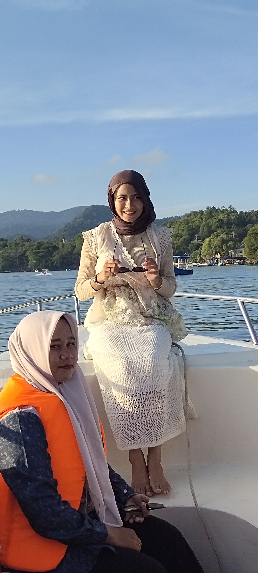 Two women are seated on the edge of a white boat. The woman in the foreground, on the left, is wearing a light purple hijab and an orange life vest over a dark blue shirt with a floral pattern. She is looking towards the camera with a neutral expression and holding a small object in her right hand. The woman in the background, on the right, is wearing a dark purple hijab and a cream-colored outfit with intricate lace detailing. She is smiling and holding sunglasses in her hands, looking slightly to her left. The boat is on a body of water with calm, blue waves. In the background, lush green hills and mountains are visible, dotted with trees and some buildings. The sky is clear and blue with a few wispy clouds. The scene appears to be set in Sabang, Indonesia, likely during a boat tour.