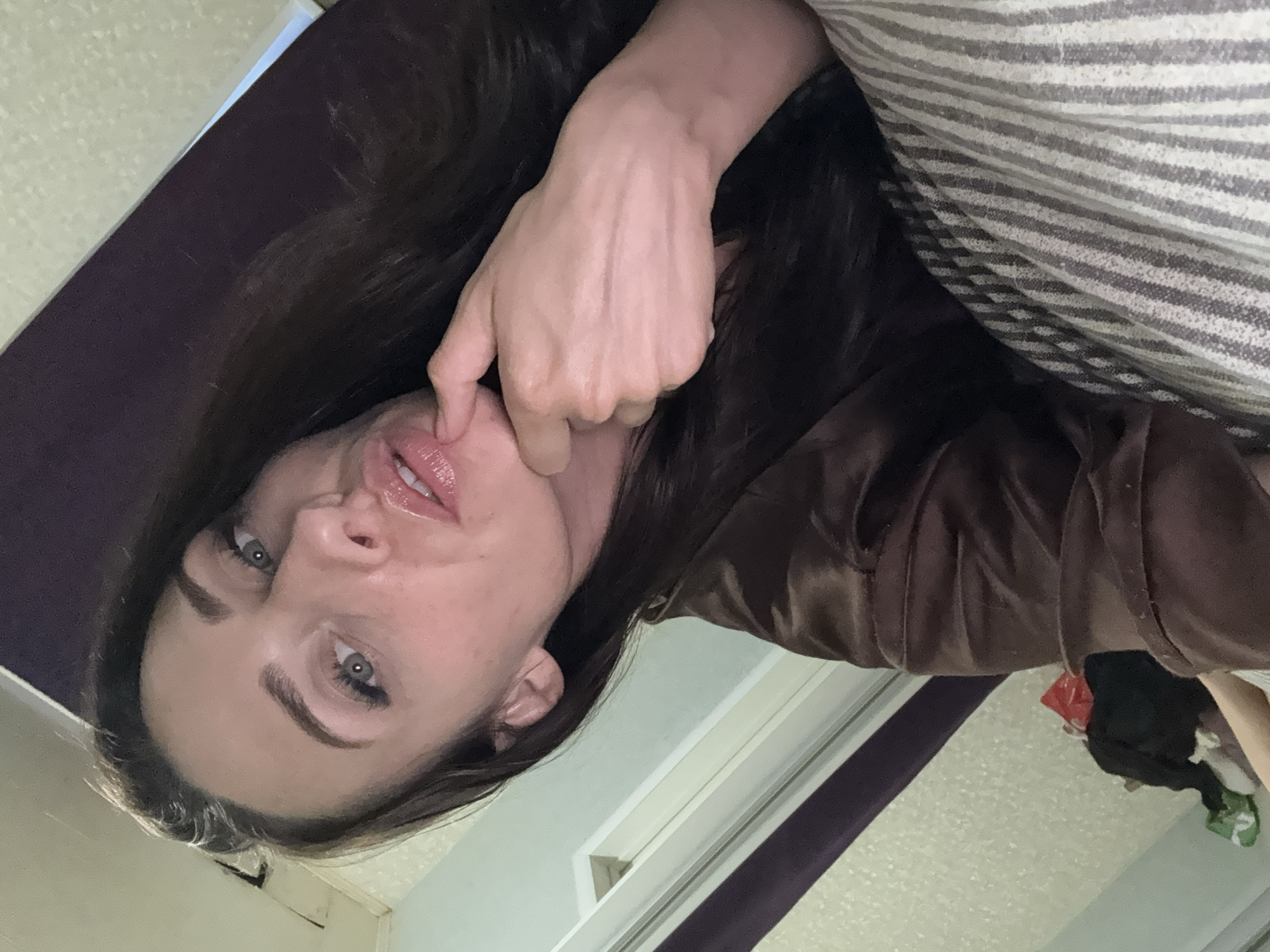 A woman is taking a selfie in a dimly lit room. She has long dark hair and is wearing a brown satin robe. Her right hand is touching her chin. In the background, there appears to be a bed with striped bedding and some items of clothing on the floor. The room also has what looks like a closet with a purple interior and a white door. The overall scene suggests a private, domestic setting.