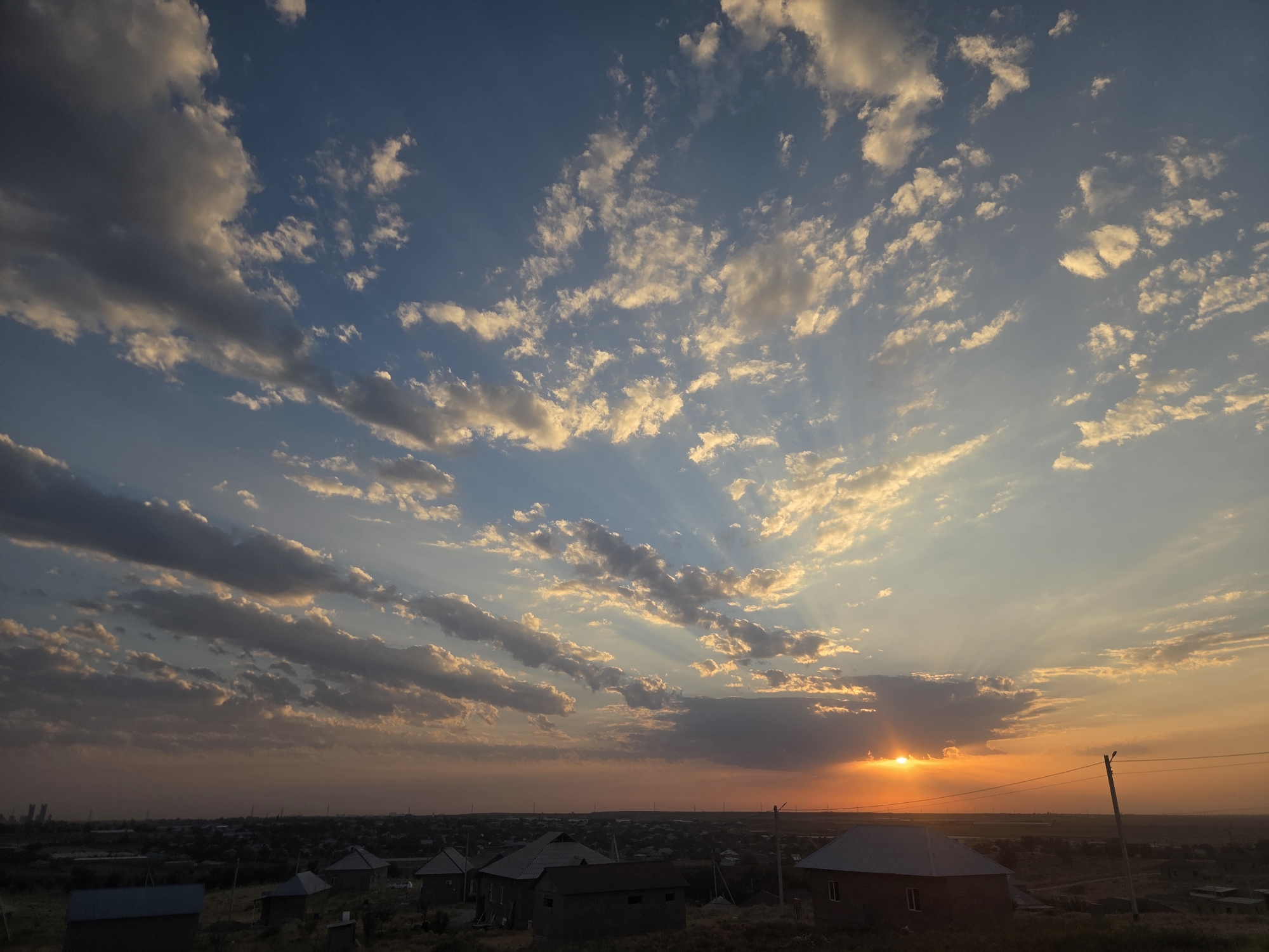 The image depicts a sunset over a semi-rural area, likely on the outskirts of Қарасу, Kazakhstan. The sky is filled with a dramatic display of clouds, illuminated by the setting sun which is partially obscured by a large cloud formation. Rays of sunlight pierce through the clouds, creating a crepuscular effect. In the foreground, there are several small residential structures with dark roofs, appearing to be simple houses or outbuildings. The landscape is somewhat arid, with dry vegetation visible around the buildings. In the distance, a hazy urban or industrial skyline can be discerned, suggesting the presence of a larger settlement beyond the immediate area. The overall impression is one of a calm, natural scene transitioning from day to night. There are no people or animals visible in the image.