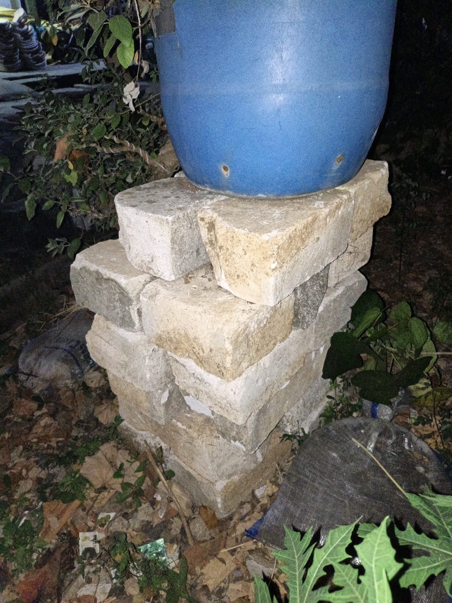A blue pot sits atop a stack of light-colored, rough-textured bricks. The bricks are unevenly stacked, creating a makeshift pedestal. The ground around the bricks is covered with dry leaves, some green foliage, and a dark, textured material that appears to be a tarp or bag. In the background, there are out-of-focus plants and shadows, suggesting an outdoor, possibly neglected, setting. There are no people visible in the image. The scene is dark, indicating it was likely taken at night or in very low light conditions. The location is Cikal, Indonesia.