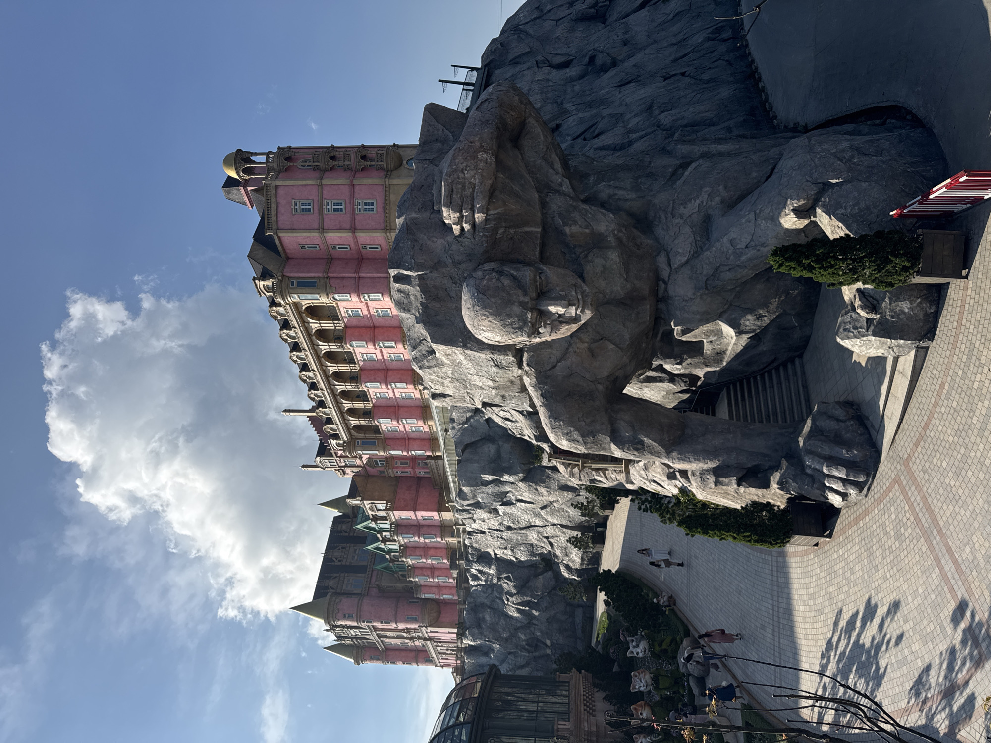 The image displays a panoramic view of Sun World Ba Na Hills, located in Da Nang, Vietnam. Dominating the upper portion is a large, multi-story building constructed in a French château architectural style. It features pink-colored walls, dark conical roofs, multiple turrets, balconies with arched openings, and dormer windows. This structure is situated atop a massive, rugged artificial rock formation.
Integrated directly into the rock formation below the building is a colossal, gray stone sculpture of a muscular man. The figure is depicted in a stooping posture, with his head facing forward and slightly downward, and one arm raised, bent at the elbow, seemingly supporting or emerging from the rock. Dark steps lead into an opening located within the lower portion of the sculpture.
At ground level, a wide, light-colored paved walkway, edged with a reddish border, curves gently to the left. Along the left side of the path, a landscaped area contains various green topiary sculptures, including several large cat heads, and a partial view of a glass-domed structure. Several individuals are present on the walkway; four women are clearly visible in the mid-ground, with two walking abreast in light attire, one in a light top and skirt, and another further back in a pink dress. Small, conical evergreen trees in dark wooden planters are spaced along the path. A red and white striped barrier is partially visible on the far right.
The sky is clear blue in the upper right, transitioning to a large, prominent white cumuliform cloud in the upper left and center. The scene is brightly illuminated by direct sunlight, casting distinct shadows on the ground.