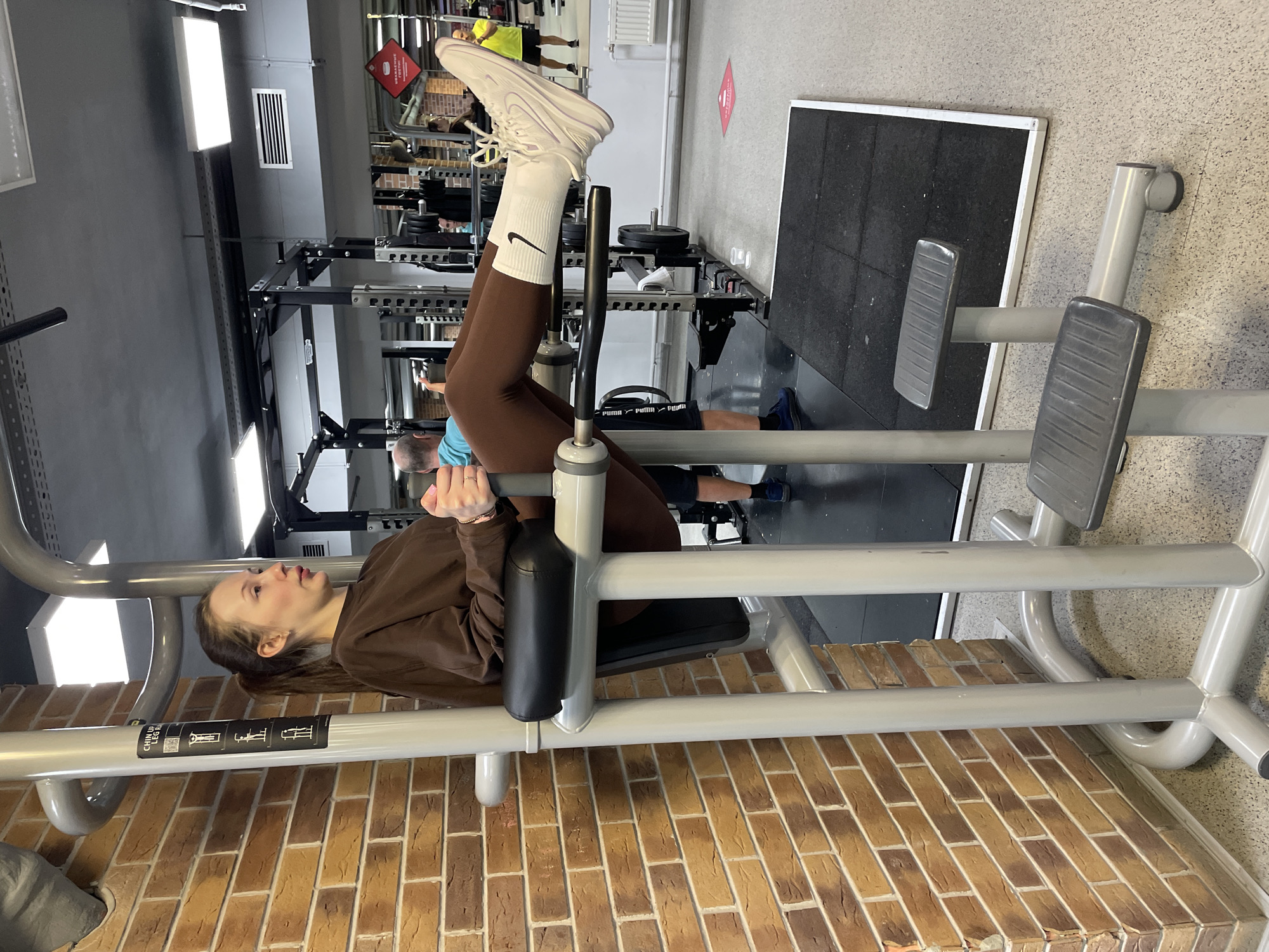 A young woman is using a chin-up and leg-raise machine in a gym in Izhevsk, Russia. She is hanging upside down, gripping the machine handles, with her legs raised and knees bent. She wears brown leggings, white socks with a Nike swoosh logo, and white sneakers. She also wears a brown long-sleeved shirt. The machine is silver with a black padded backrest. The gym has other weightlifting equipment in the background, including a squat rack and various machines. The floor near the leg-raise machine is made of brick-like material.