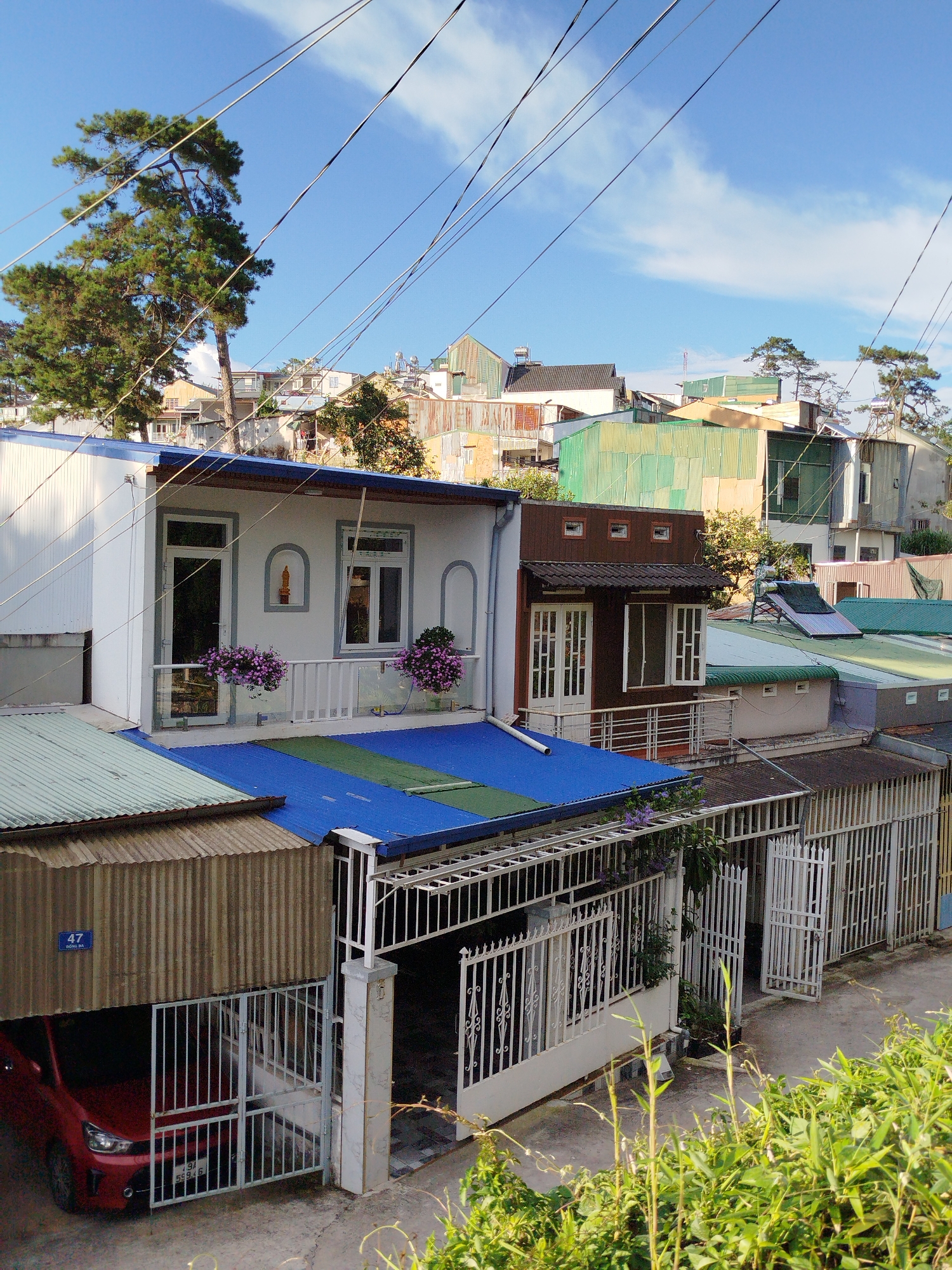 The image depicts a residential street scene in Da Lat, Vietnam, characterized by its hilly terrain and a dense collection of houses with corrugated metal roofs. In the foreground, a red car is parked under a covered carport. A white metal gate, partially open, leads to a paved area. The houses are multi-story structures with varying architectural styles, some featuring balconies with potted plants. Power lines crisscross the sky, typical of urban Vietnamese settings. Tall pine trees are visible, hinting at Da Lat's mountainous location and cooler climate. The overall impression is one of a densely populated urban area with a blend of older and more modern construction.