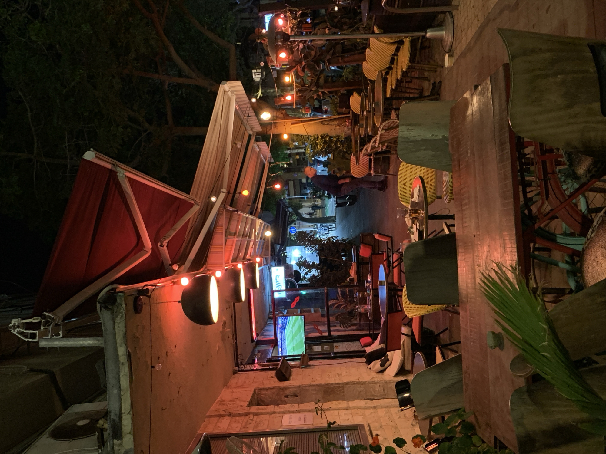 This nighttime image captures an inviting outdoor dining or bar area, likely in Tel Aviv, Israel. The scene is illuminated by numerous artificial light sources, including string lights, wall-mounted lamps with black shades, and bare bulbs, casting a warm, ambient glow across the space.
The setting appears to be a narrow alleyway or pedestrian street converted into an al fresco establishment. It features various seating arrangements, including tables with dark tops and chairs, many of which have distinctive yellow woven backs. Red and striped awnings or large umbrellas provide cover over some of the seating areas. Green foliage from potted plants and trees is visible throughout, adding to the atmosphere.
In the mid-ground, a man wearing a dark top is seen standing near tables, possibly a patron or staff member. On the left side of the image, a television screen displays what appears to be a soccer match, indicating entertainment for guests. Architectural elements include stone walls, wooden structures that might serve as bars or counters, and visible air conditioning units.
Several signs with Hebrew text are discernible, confirming the location context. A vertical sign above the TV screen appears to read "קליאו" (Kleo or Cleo), and other smaller signs on the wall and a document on the ground also display Hebrew characters. A blue circular sign is visible further down the path. The overall impression is one of a lively, relaxed, and social evening atmosphere.