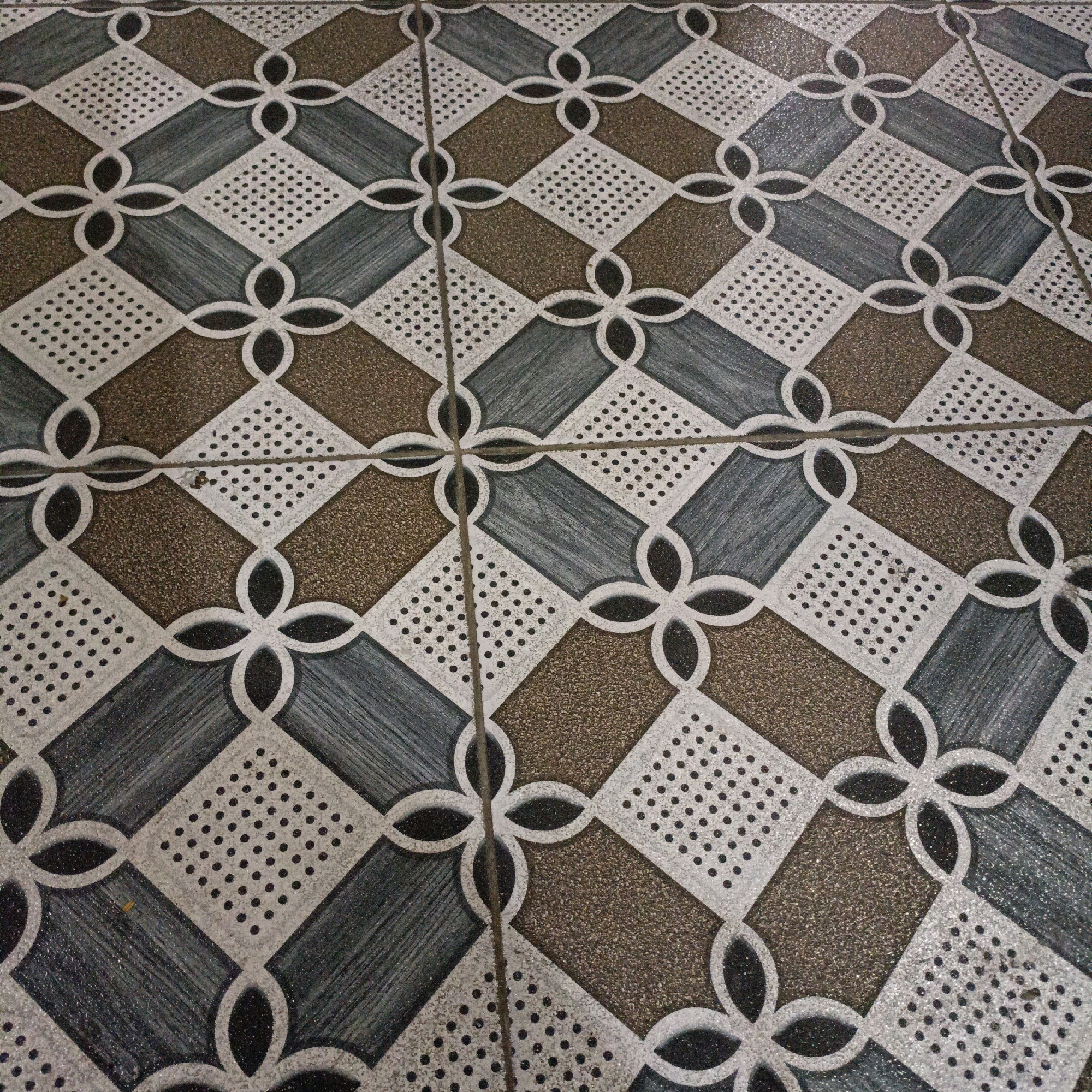 The image shows a close-up, angled view of a tiled floor with a geometric pattern. The tiles are arranged in a diamond grid, with each diamond containing a smaller, dotted diamond shape in its center. Interconnecting floral or clover-like shapes in white outline are present at the intersection of the larger diamond tiles. The larger diamonds themselves are patterned with alternating colors and textures: brown speckled, dark gray with fine vertical lines, and white with fine black dots. The floor appears to be indoors, possibly in a public space given the durable tile material. There is no indication of the specific location within Salatiga, Indonesia, but the tile pattern suggests it could be a commercial building, a traditional market, or a public facility. No people, actions, or interactions are visible.