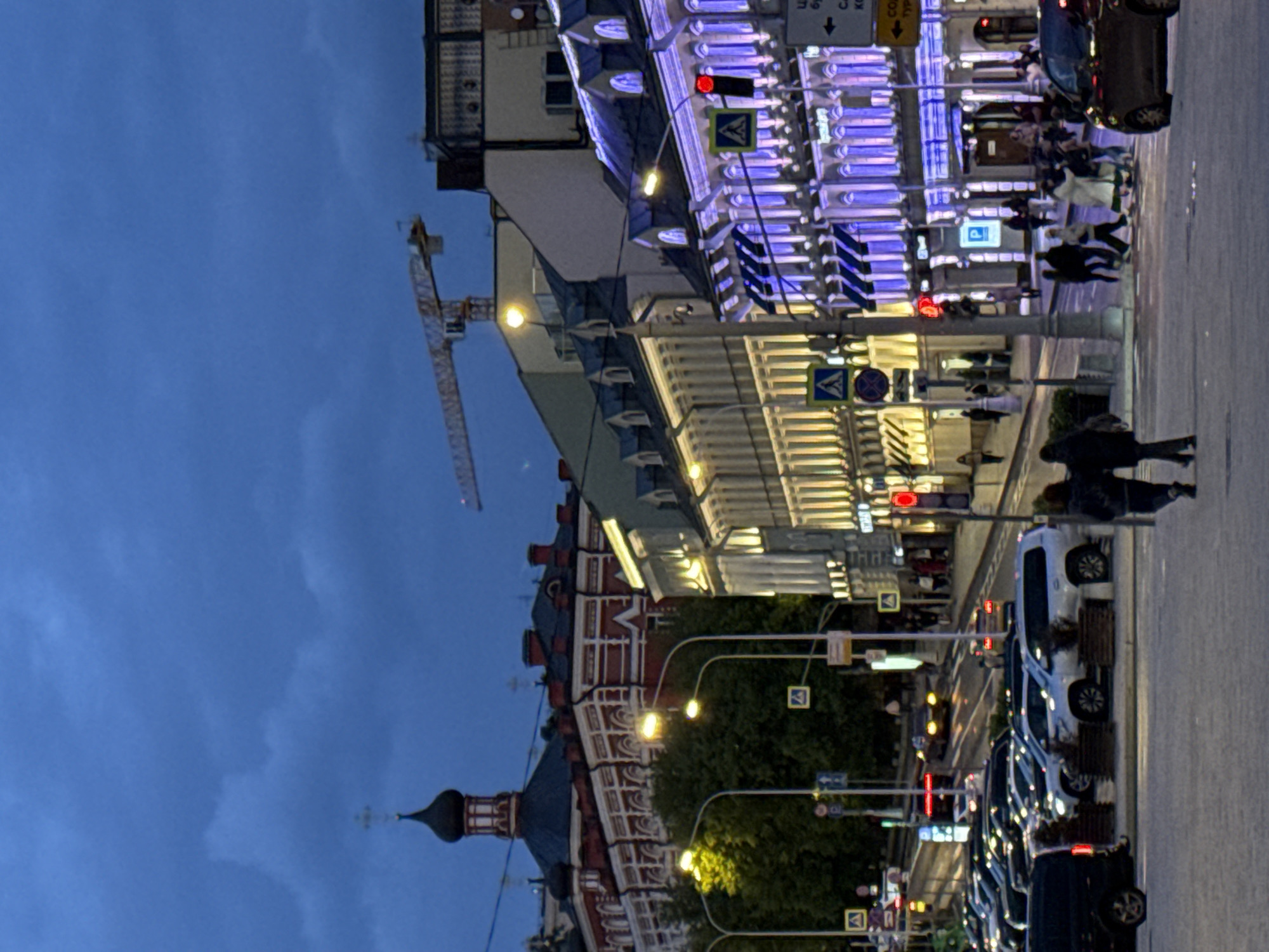 The image depicts a street scene in Moscow, Russia, at dusk or night. The view is oriented sideways. On the right side, a line of parked cars stretches along a sidewalk where pedestrians are walking. Streetlights illuminate the scene. Buildings are visible on both sides of the street, including one with a distinctive spire and ornate red and white facades. Another building is lit with blue and white lights. In the distance, there is a construction crane visible. Traffic signals are present, some displaying red lights. The sky is a deep blue color.