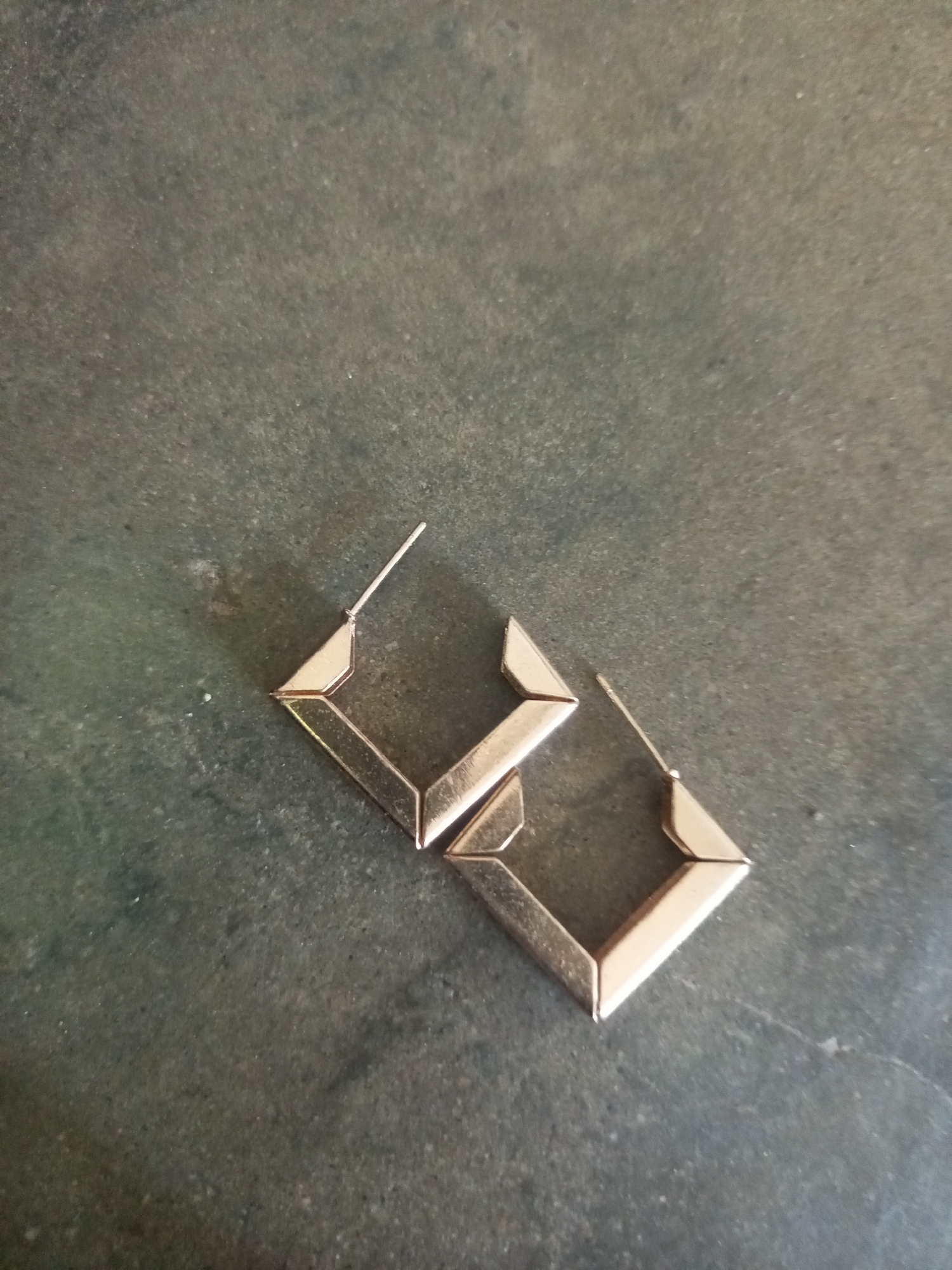 A pair of gold-colored, geometric earrings are displayed on a dark, textured surface. The earrings are shaped like open squares with pointed corners, giving them a diamond-like appearance. One earring is positioned slightly above and to the left of the other, with their post backs visible. The surface appears to be a stone or concrete material with a mottled gray and green coloration. No people are present, and the scene is static. The image does not provide any information about the location in La Tagua, Venezuela.