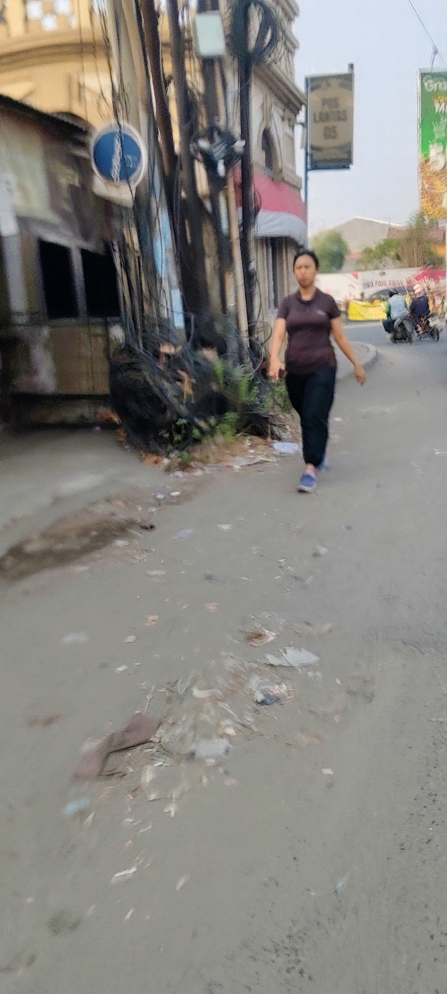 A medium shot captures an Indonesian woman walking on a dirty, uneven asphalt road in Pasirsari, Indonesia. She is wearing a dark maroon, short-sleeved shirt and dark pants, with blue sneakers. Her posture indicates she is walking forward, possibly with her phone in her left hand. The environment is cluttered with visible piles of debris and trash on the roadside, and an array of electrical wires hangs overhead, connected to a pole in the mid-ground. Behind the woman, a building with a light-colored facade and windows is partially visible, along with signs. In the background, other individuals are seen on motorcycles. The lighting suggests it is daytime, possibly late afternoon, given the soft light.