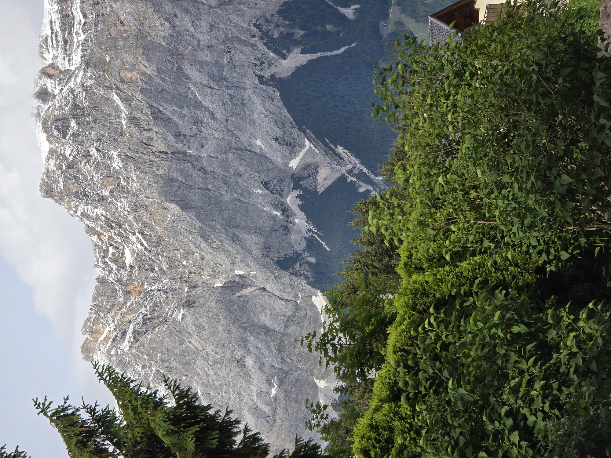 This is a vertical, medium-shot photograph of a rocky, snow-dusted mountain range looming behind a foreground of lush green trees and bushes. A portion of a building with a tiled roof and a balcony is visible to the right, peeking through the foliage. The mountain's grey rock face is deeply carved and textured, with patches of snow clinging to its upper reaches and in crevices. The sky above is overcast, with a few wispy clouds. The scene captures a view that could be seen from a residential area in Lermoos, Austria, given the presence of the building and the characteristic alpine landscape.