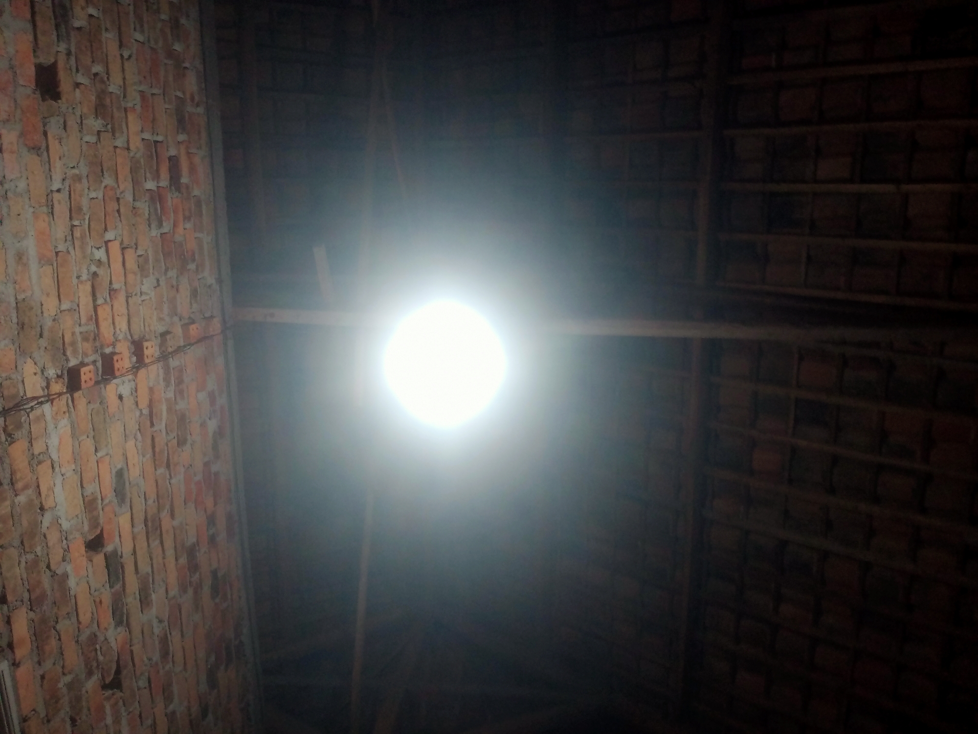 An interior shot of a brick wall and a thatched roof is captured, with a bright light source dominating the center of the frame. The brick wall, visible on the left, is constructed from reddish-brown bricks with visible mortar. The thatched roof, occupying the right side of the image, appears dark and textured. The light source is a brilliant, blown-out white circle, suggesting it is a strong lamp or the sun. There are no people or specific actions depicted in the image. The environment appears to be a rudimentary structure, potentially under construction or in a state of disrepair, located within the city of Nusa Agung, Indonesia.