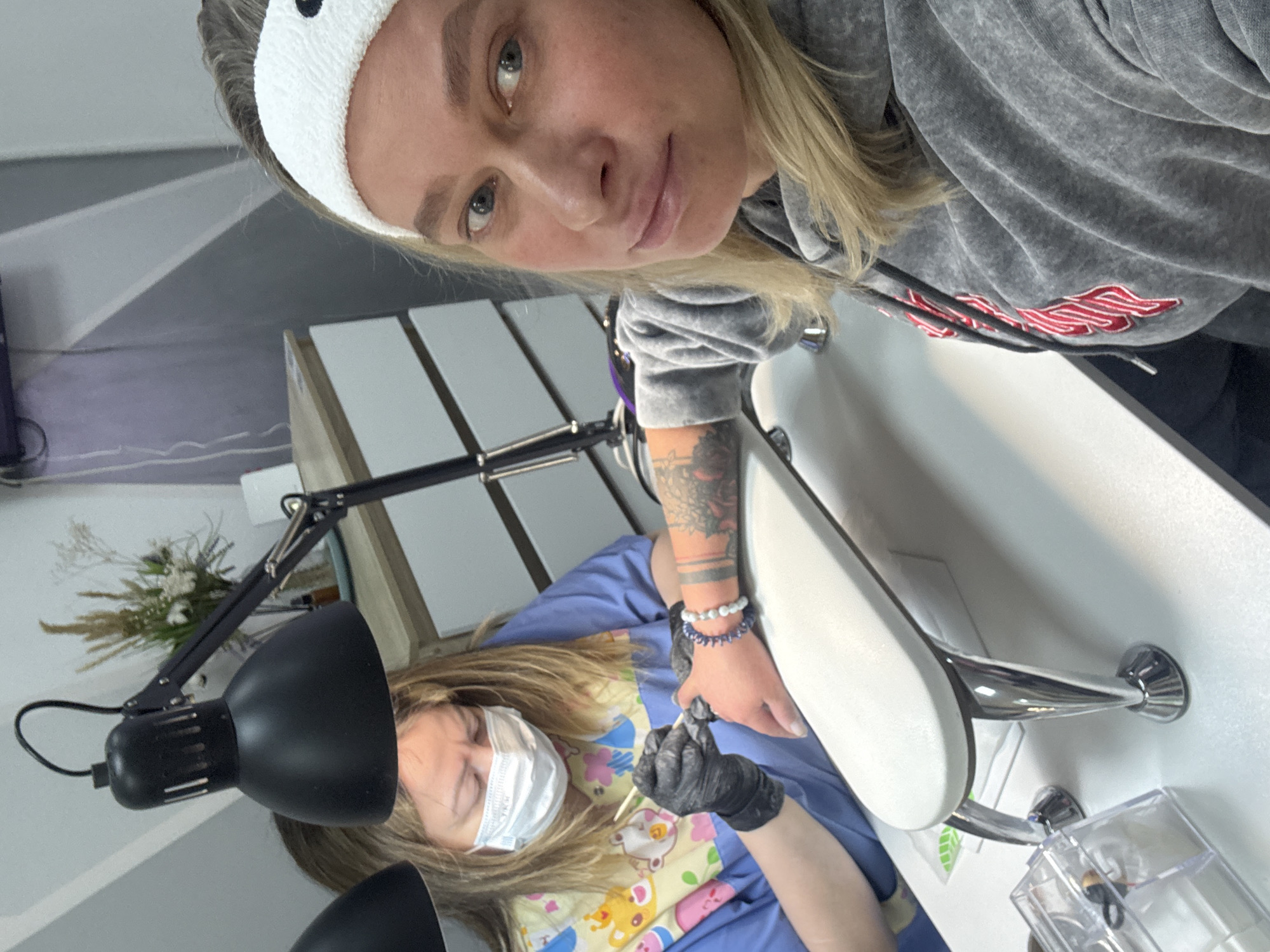 A woman with blond hair and a white headband is taking a selfie while at a manicure salon in Perm, Russia. The selfie includes a manicurist wearing a blue uniform and a white surgical mask. The manicurist is seated under an adjustable black lamp, working on the fingernails of a client with black gloves. The manicure station includes a white armrest, a sink, and a small clear container for tools. A tattoo is visible on the client’s forearm and wrists. A bouquet of dried flowers sits in the background.