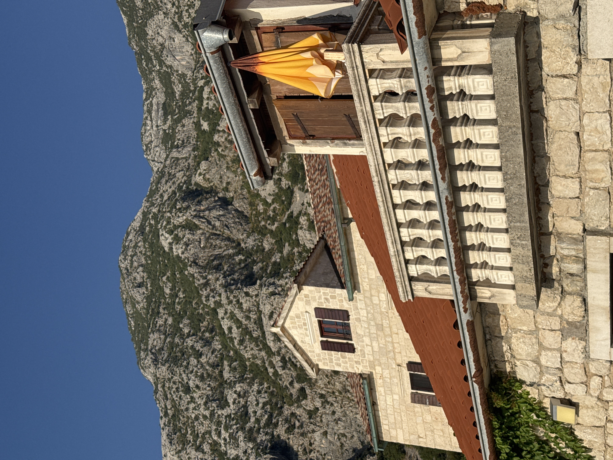 In Dobrota, Montenegro, a close-up, eye-level shot captures the facade of a stone building featuring a red-tiled roof and a decorative balustrade. Above, a rocky mountain slopes steeply against a clear blue sky, with sparse green vegetation clinging to its surface. A bright yellow umbrella, partially furled, is affixed to the exterior wall of a building, positioned on a balcony with wooden shutters. The scene is bathed in natural daylight, casting distinct shadows. The architectural style suggests a historic European setting.
