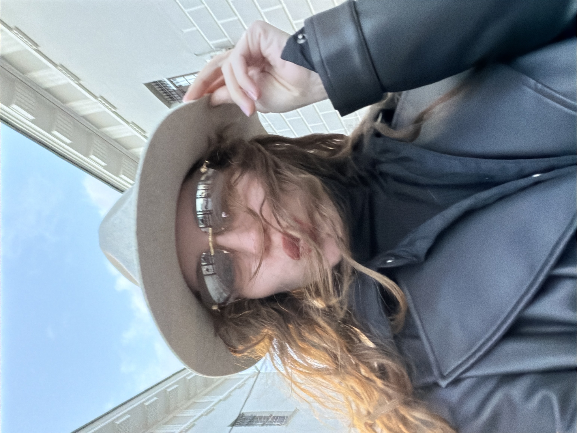 The image presents a low-angle, close-up shot of a person outdoors in Moscow, Russia, on a bright and sunny day. The subject, likely female, is captured from below, looking downwards towards the camera. She wears a light-colored, fedora-style hat and distinctive sunglasses with reflective, segmented metallic frames, partially obscuring her face.
Her long, wavy hair, in shades of brown and auburn, cascades around her face and is illuminated by the sunlight, creating bright highlights. She is dressed in a dark, possibly leather or faux leather, coat or jacket, which features a prominent collar and visible buttons on the sleeve cuffs. One hand, with subtle nail polish, is raised near her temple, suggesting she might be adjusting her hat or hair.
The background reveals a clear blue sky and sections of a light-colored building with architectural details such as ledges and window frames, indicating an urban environment. The lighting suggests it is daytime. There is no visible text in the image.